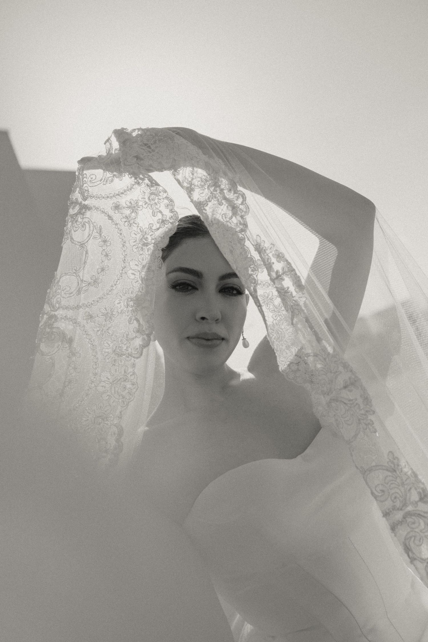 Black and white bridal portrait of elegant bride lifting lace veil, soft light creating dreamy wedding photography.