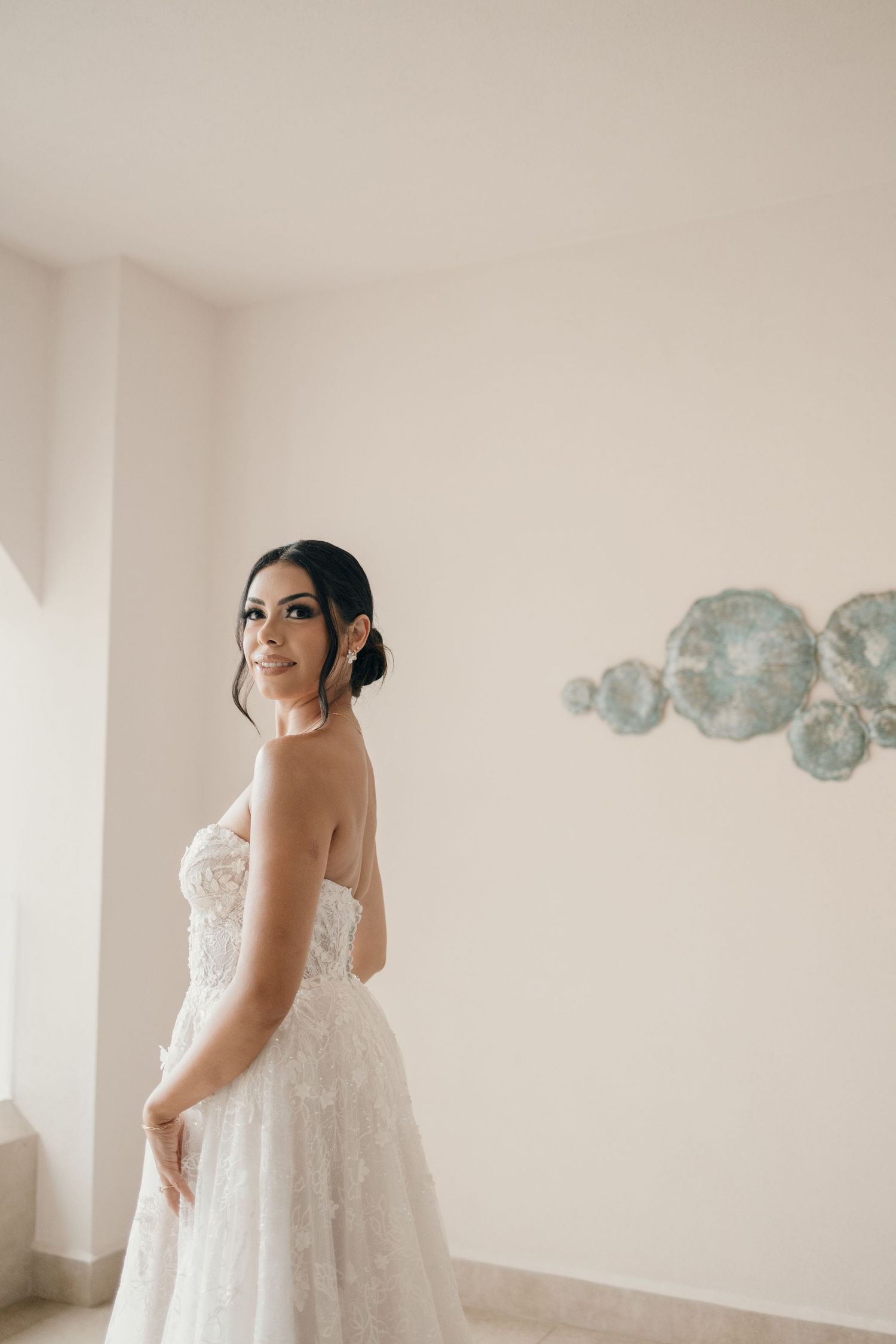 A bride in a white lace wedding dress poses in front of a light wall with decorative blue plates mounted as decor.