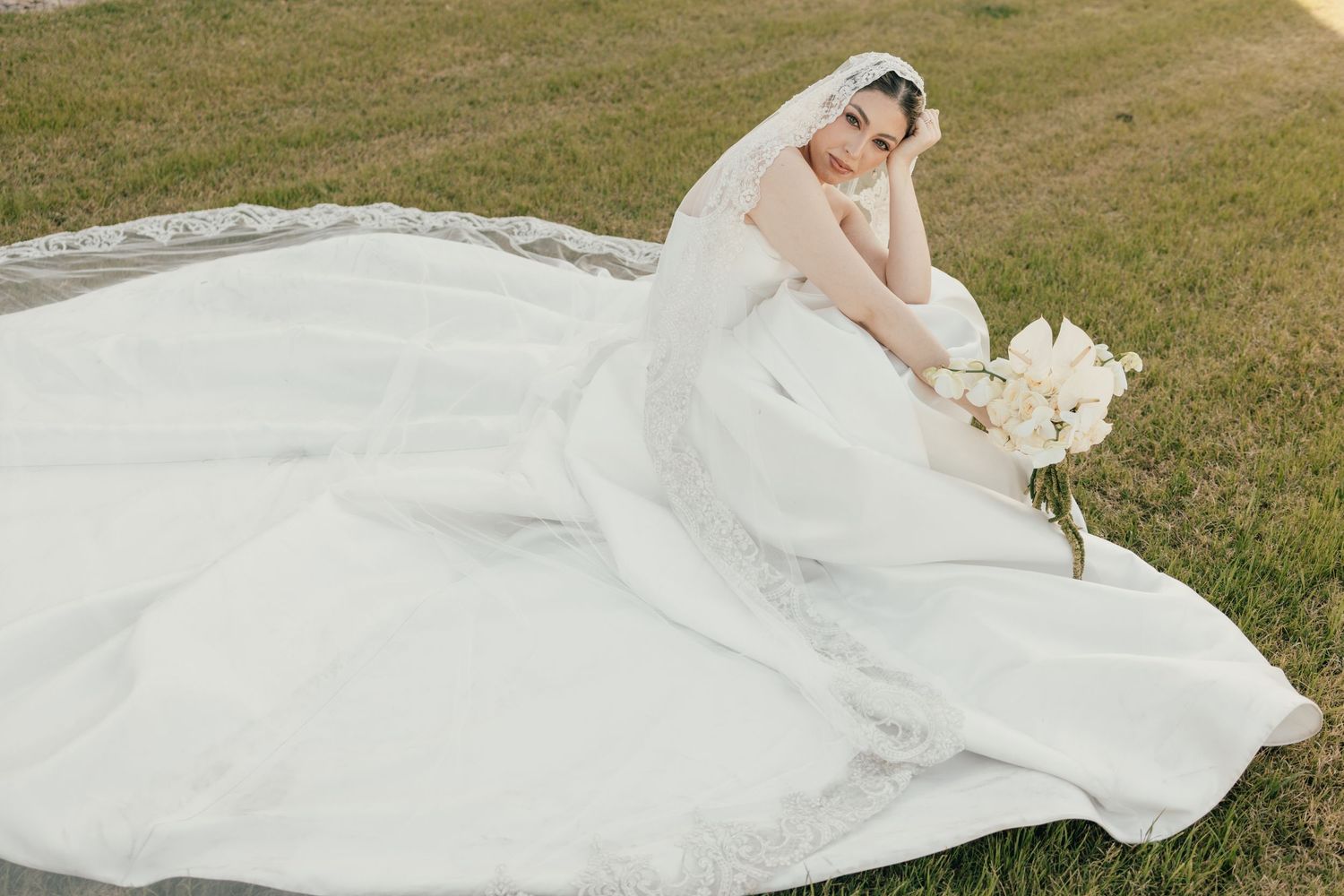 Bride in a white lace wedding gown with long veil sits on grass holding a bouquet of white flowers.