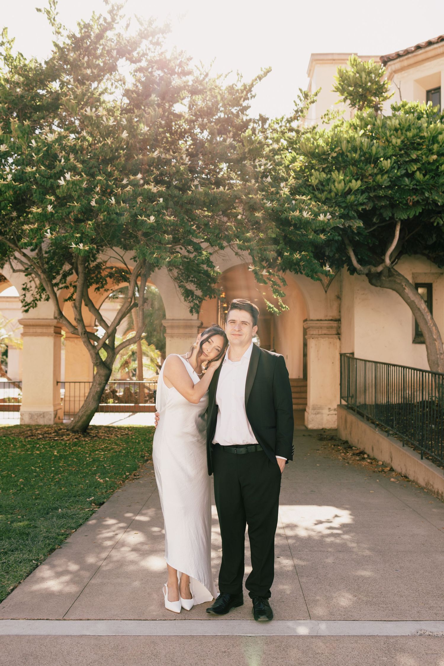 A couple takes romantic wedding photos along a tree-lined walkway with Mediterranean architecture.