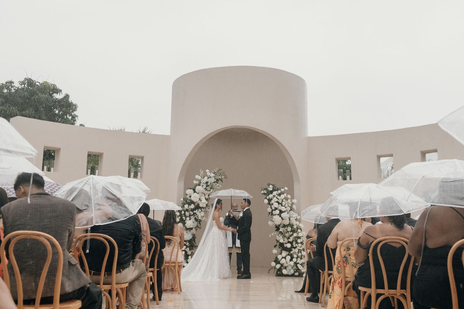 Outdoor wedding ceremony at a modern white venue with guests holding clear umbrellas on a rainy day, floral arch backdrop.