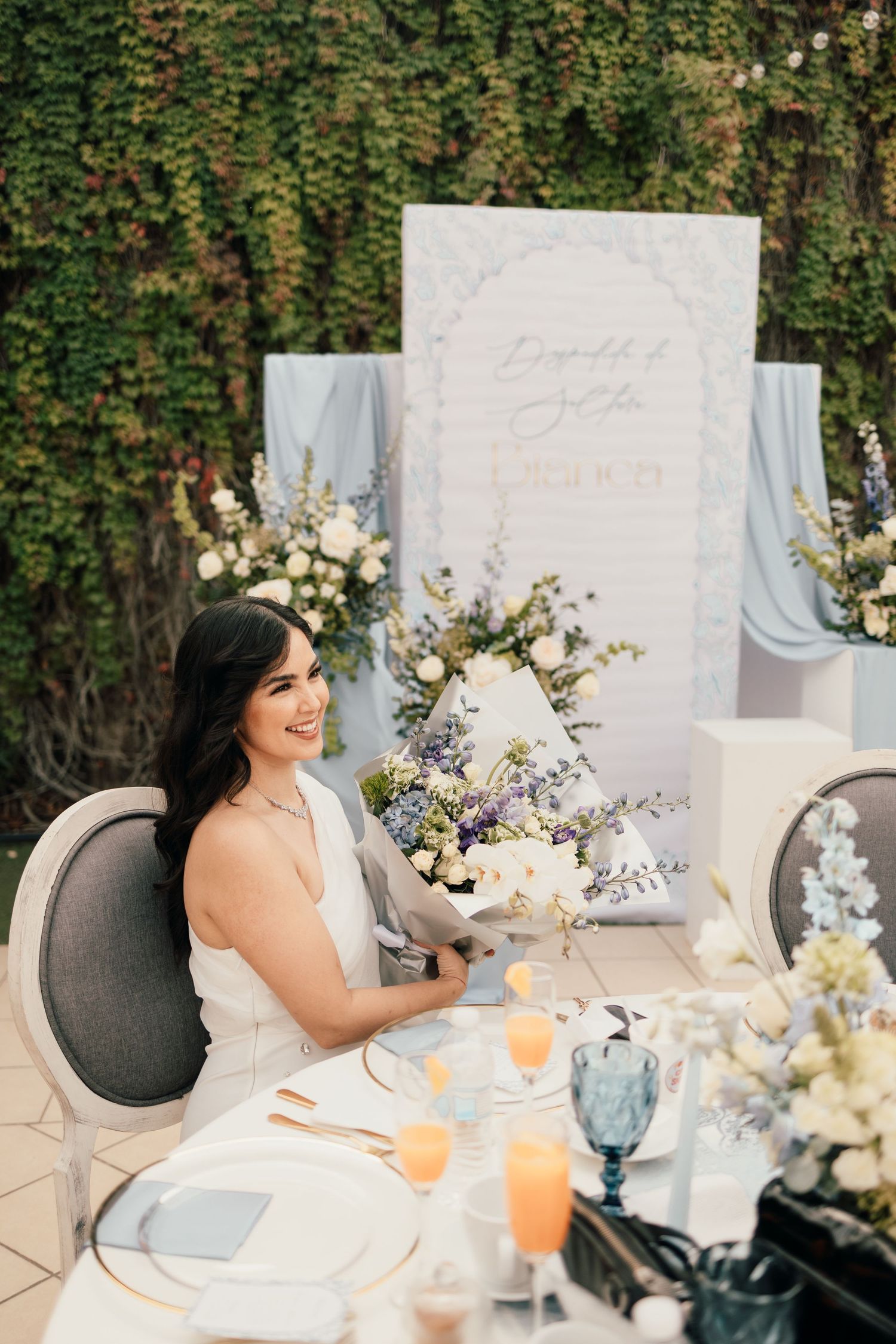 Elegant outdoor table setting with white floral arrangements and blue glassware against a lush green hedge backdrop.