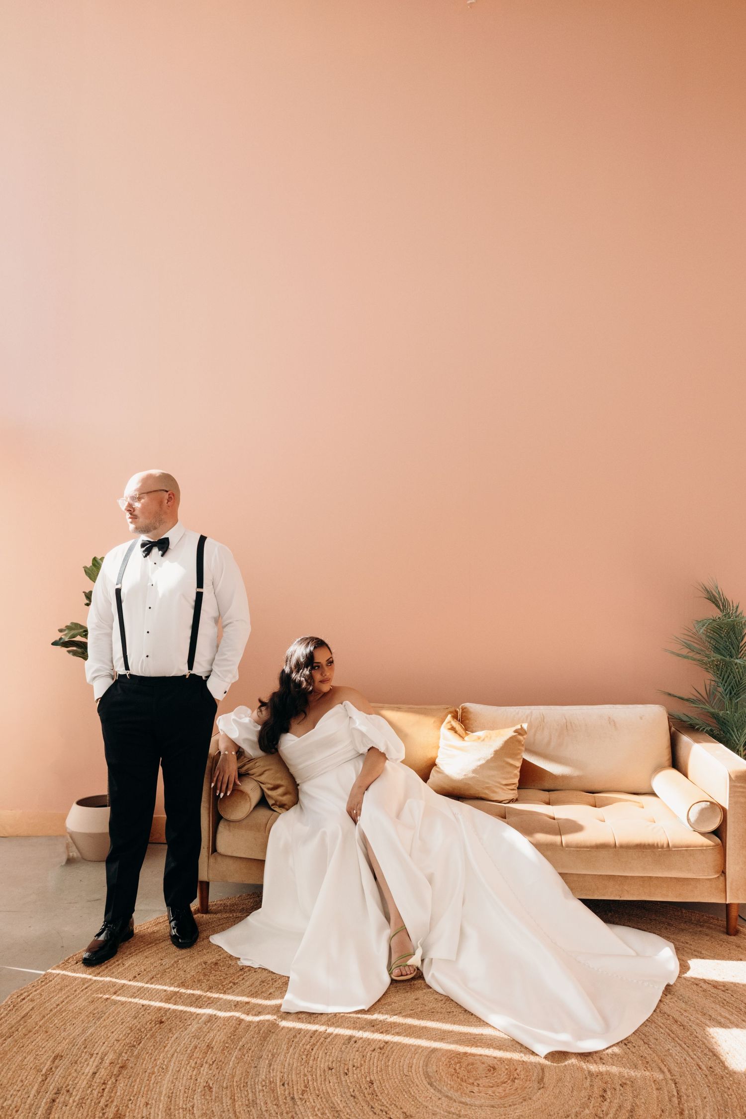 Bride in white gown reclines on beige sofa while groom in suspenders and bow tie stands beside peach wall.