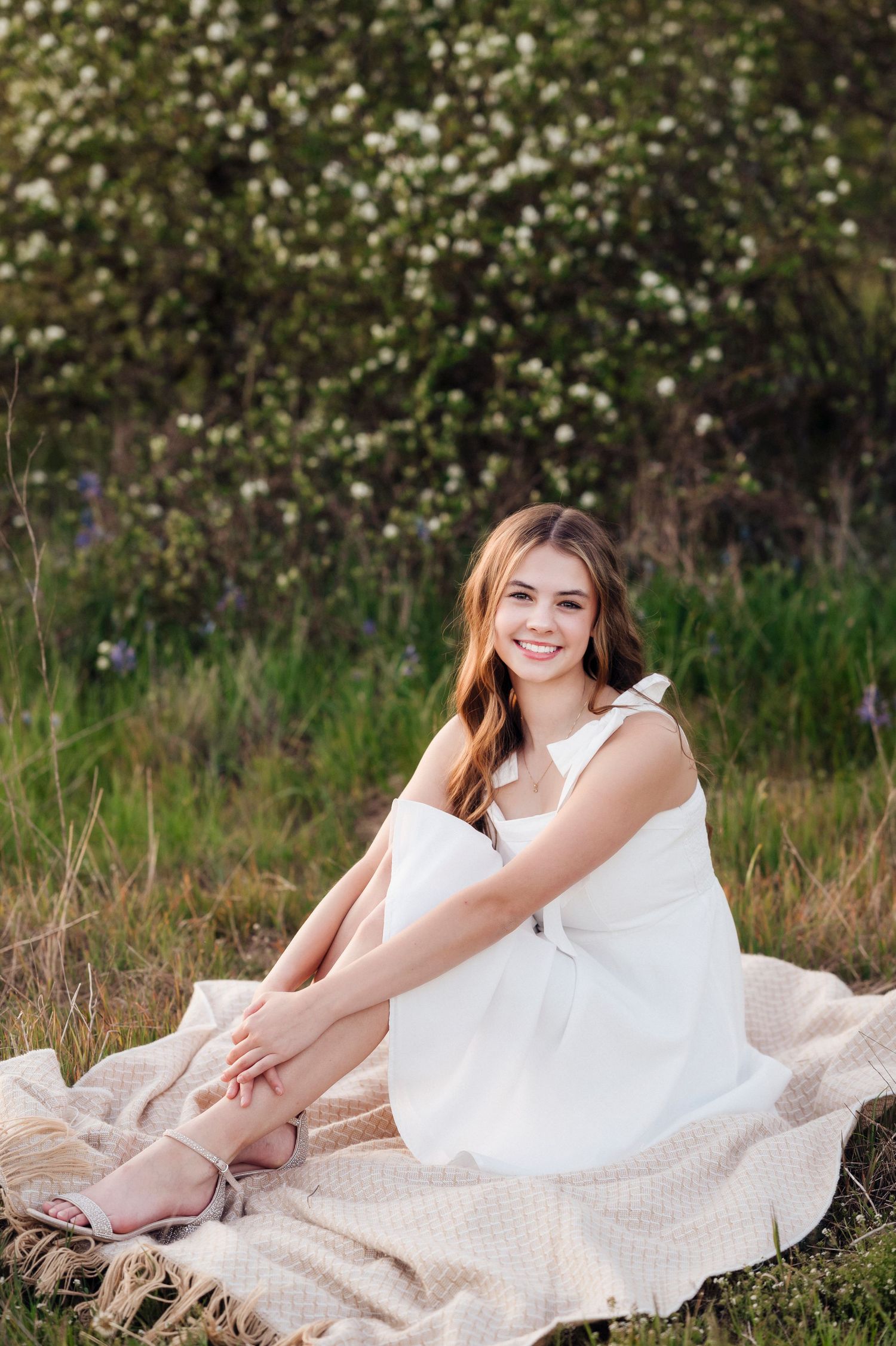 A person in a white summer dress relaxes on a blanket in a sunny meadow with blooming flowers in the background.