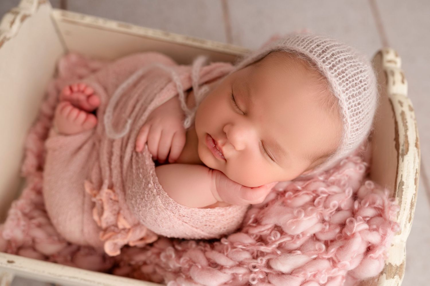 Newborn baby peacefully sleeping in white basket with pink blanket and knitted bonnet.