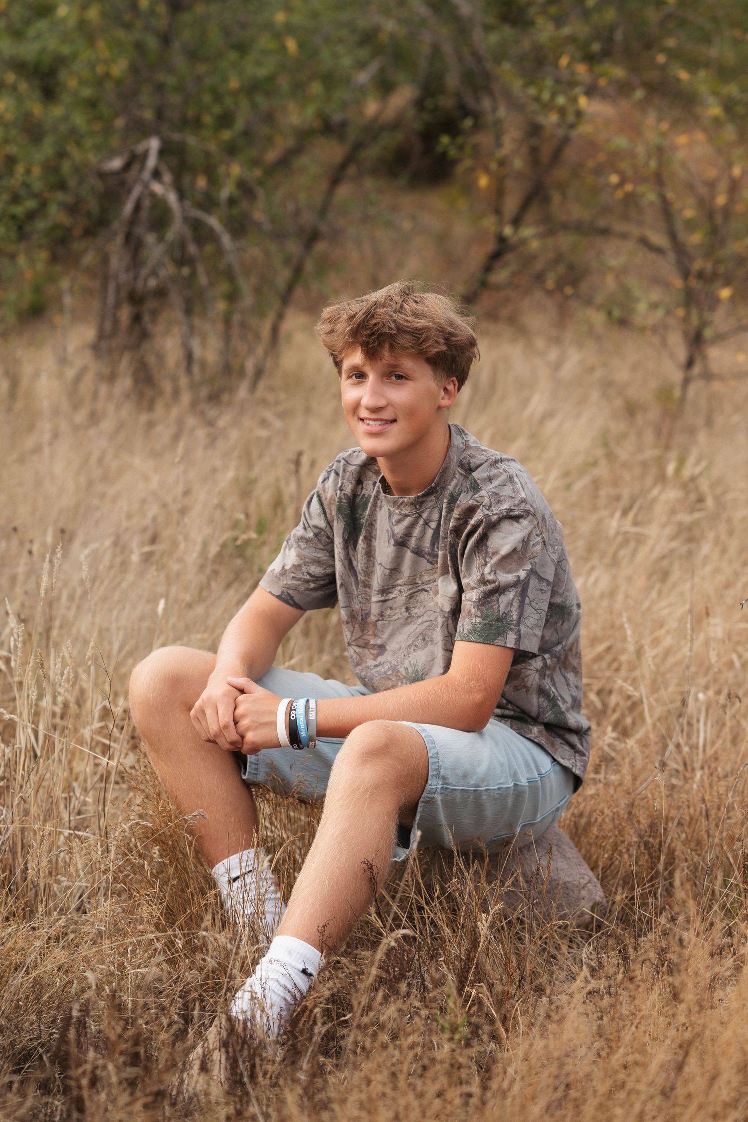 A person sits casually in a field of tall dry grass wearing denim shorts and a gray t-shirt on a sunny afternoon.
