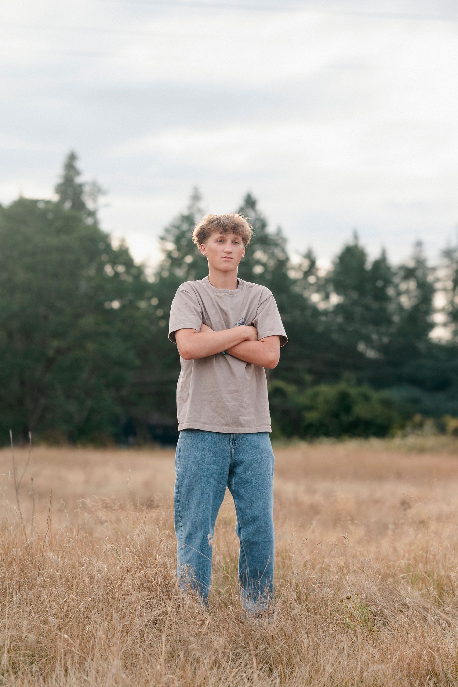Person standing in tall dry grass field with trees in background wearing grey t-shirt and jeans.