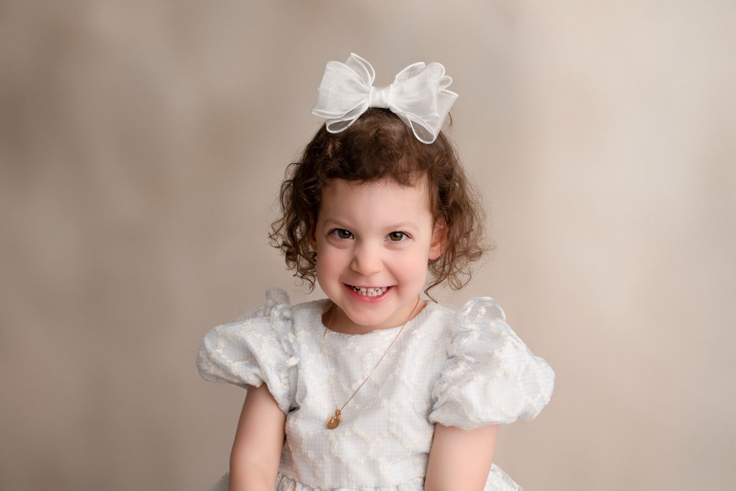 A toddler wearing a white vintage-style dress with puffy sleeves poses against a neutral beige background.