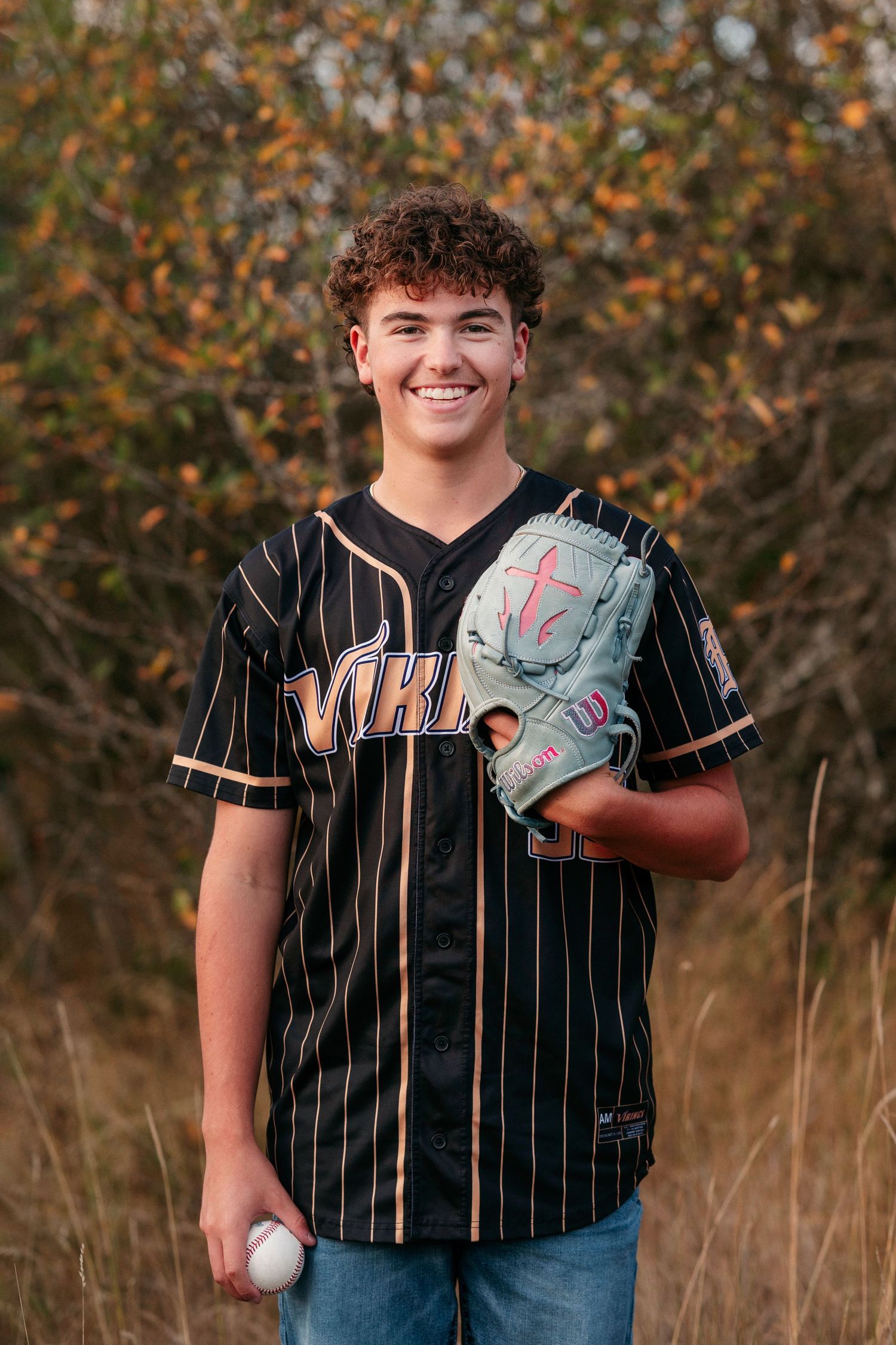 Baseball player wearing pinstriped black jersey poses with glove in autumn field with orange foliage in background.