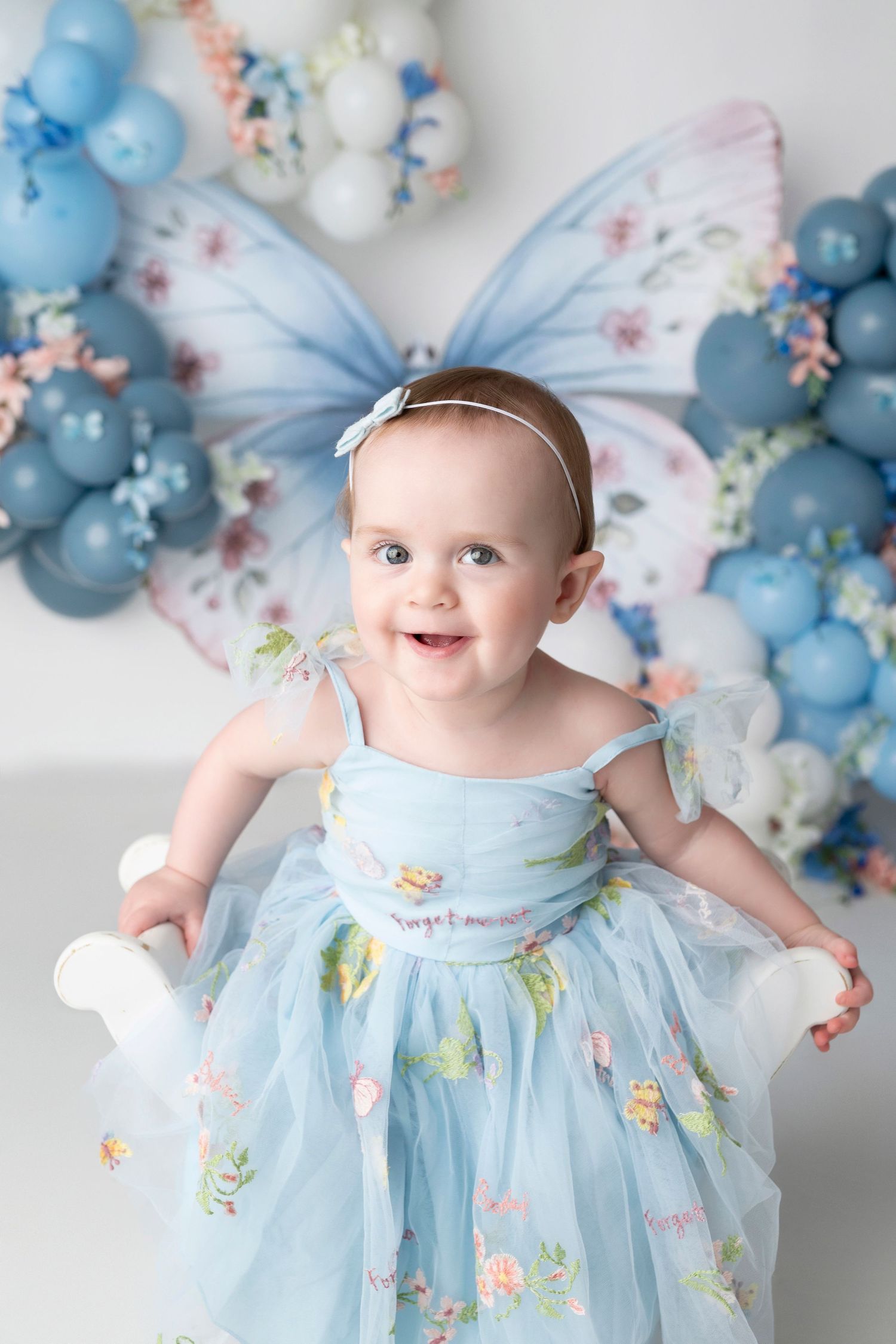 Smiling baby wearing a blue floral dress sits in front of a whimsical balloon arch with butterflies.