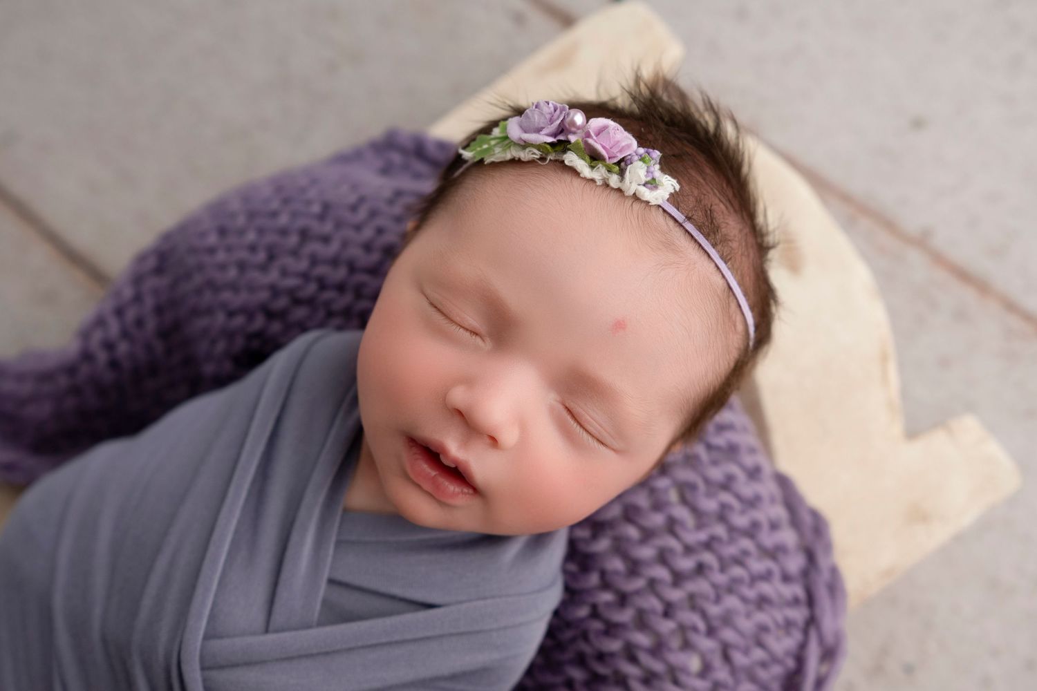 Sleeping newborn wrapped in gray swaddle wearing floral headband on purple knit blanket.