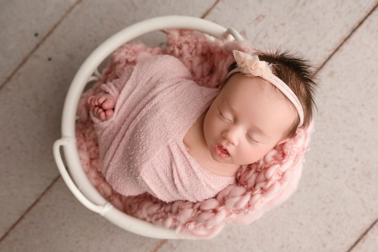 Newborn baby sleeping peacefully in white bowl with pink blanket on wooden floor.