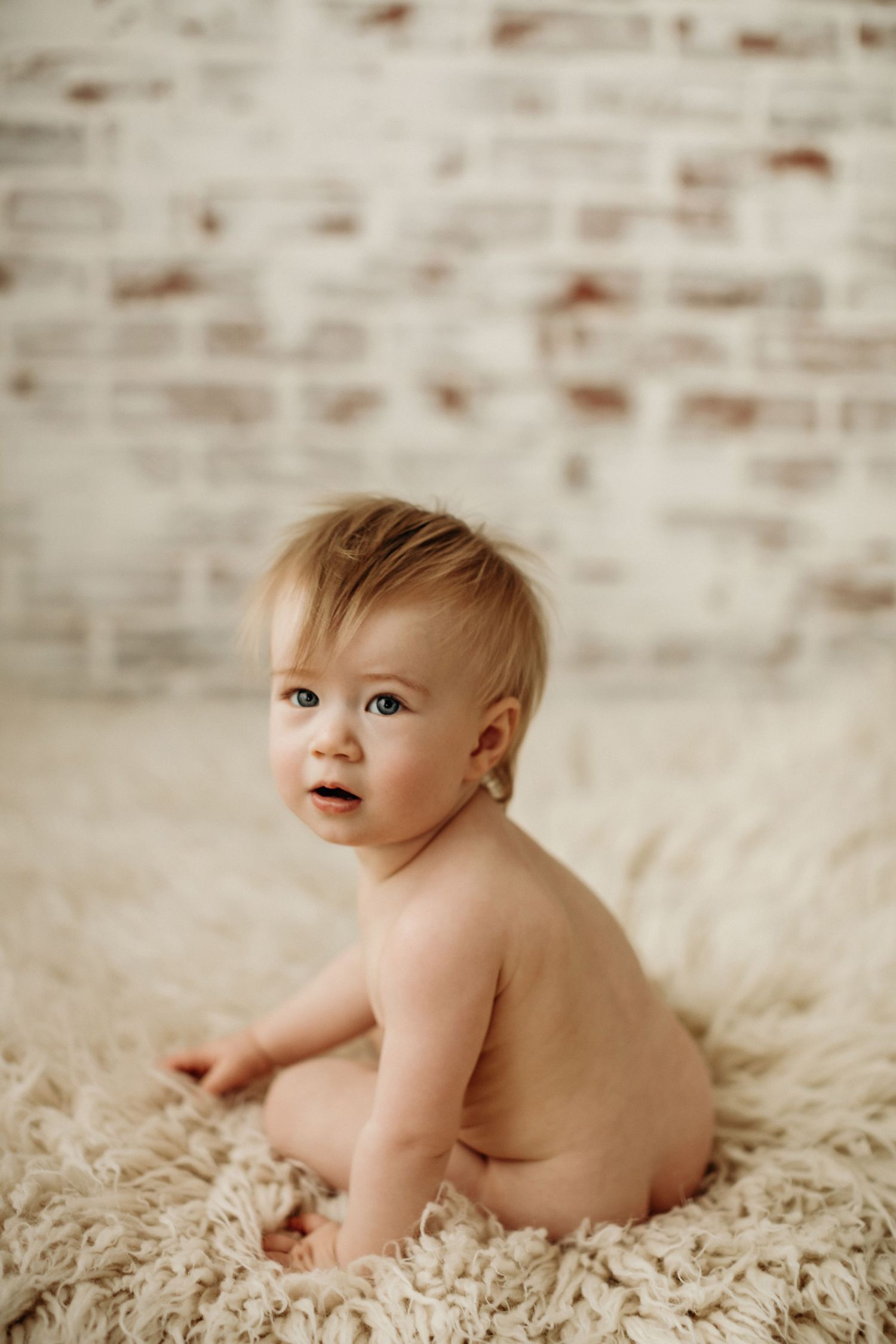 Baby sitting on cream-colored shag rug during professional portrait session against white brick wall background.