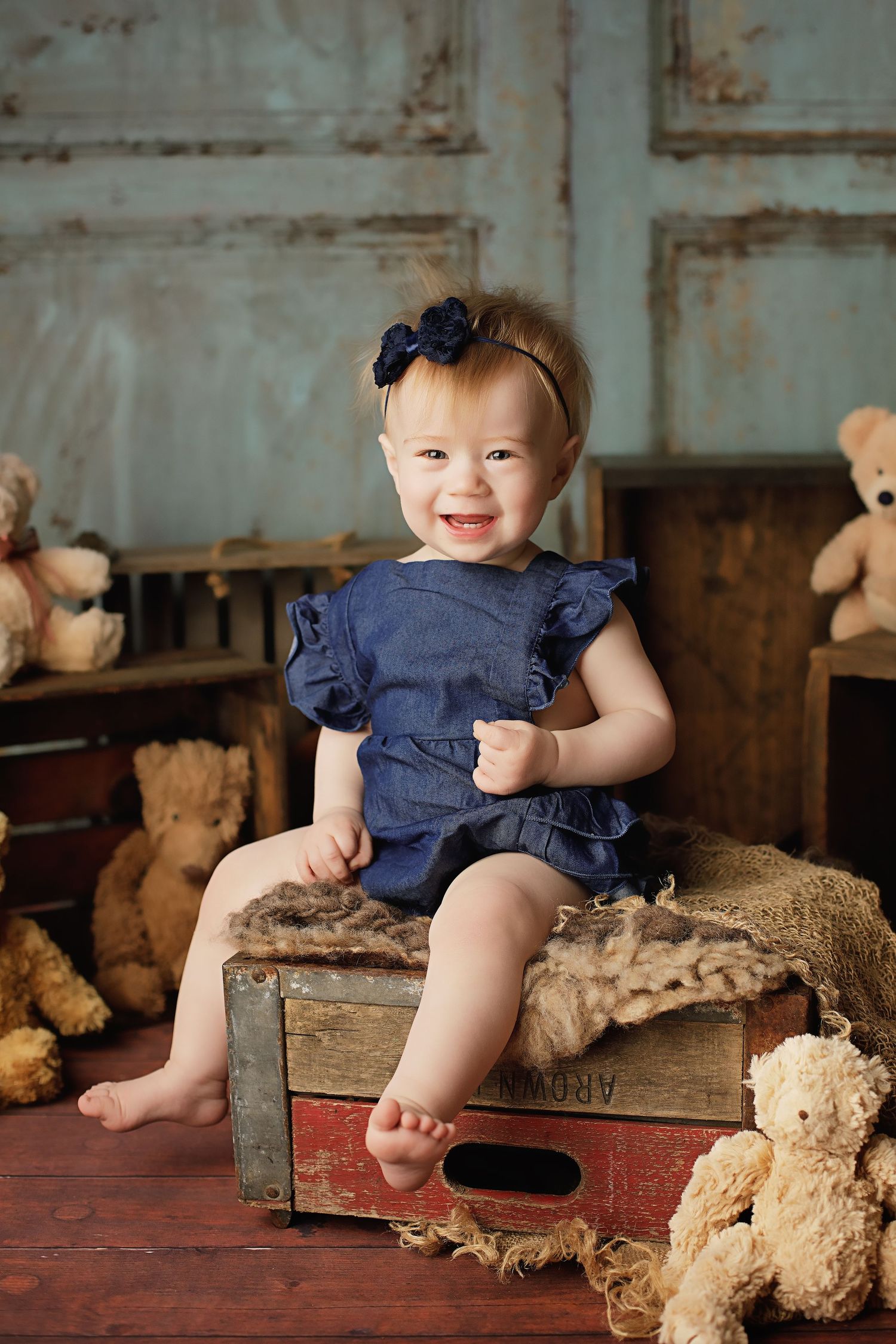 Baby wearing navy blue ruffle romper sits on vintage wooden crate surrounded by teddy bears in rustic setting.