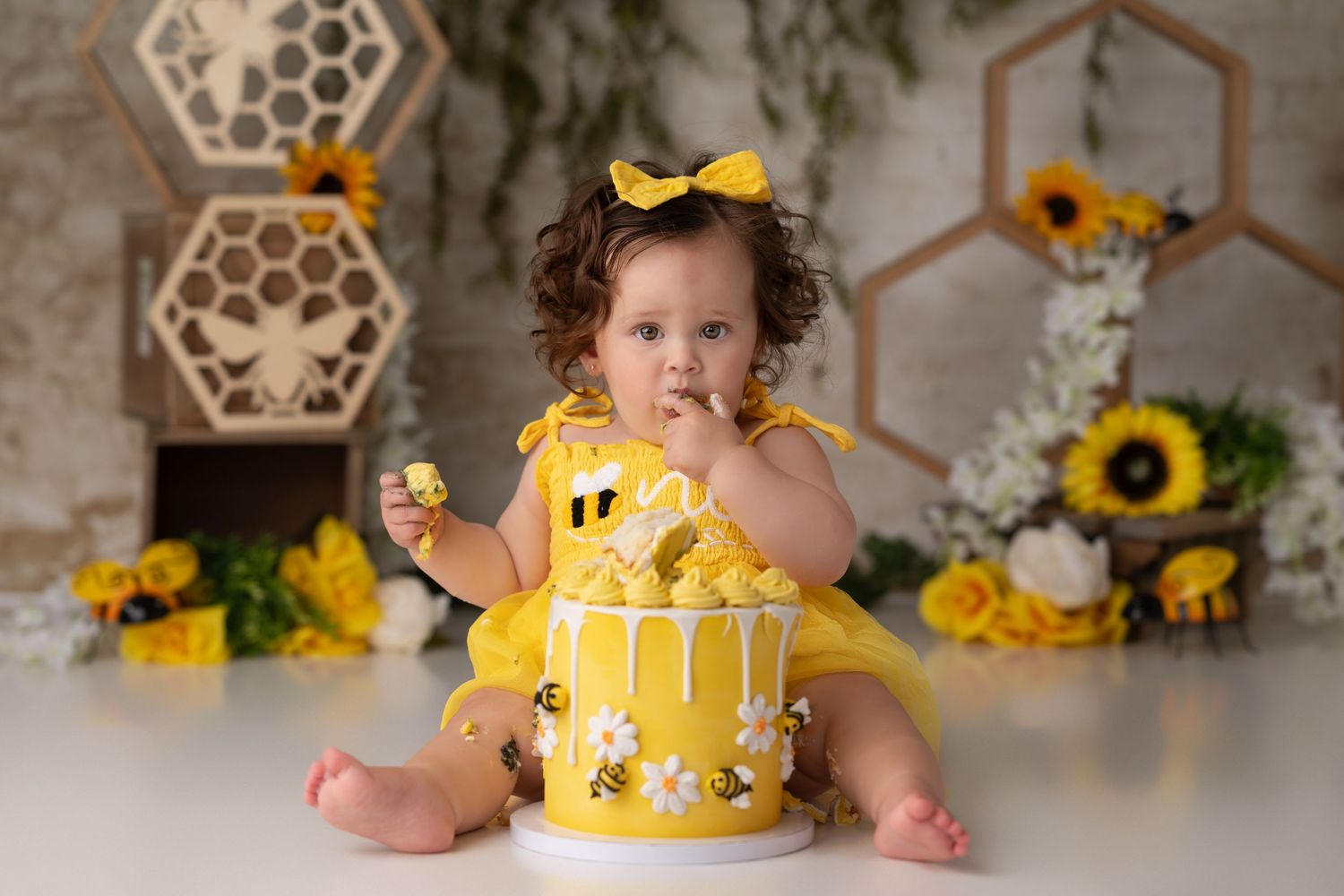 Baby girl in yellow dress smashing a bee-themed first birthday cake, surrounded by sunflowers and honeycomb decorations.