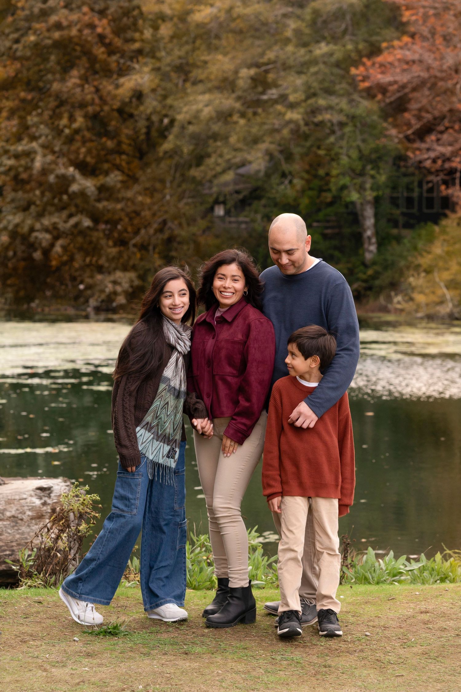 A family group stands together by a serene lake surrounded by autumn foliage during a casual outdoor photo session.