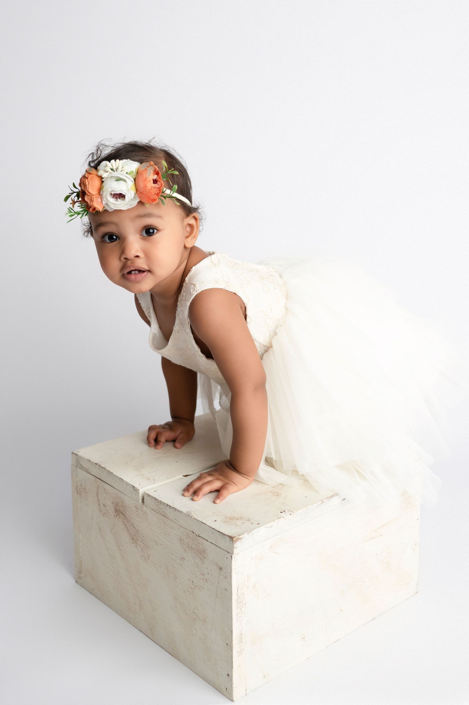 A toddler girl in a white tulle dress and floral crown poses on a white wooden box in a studio.