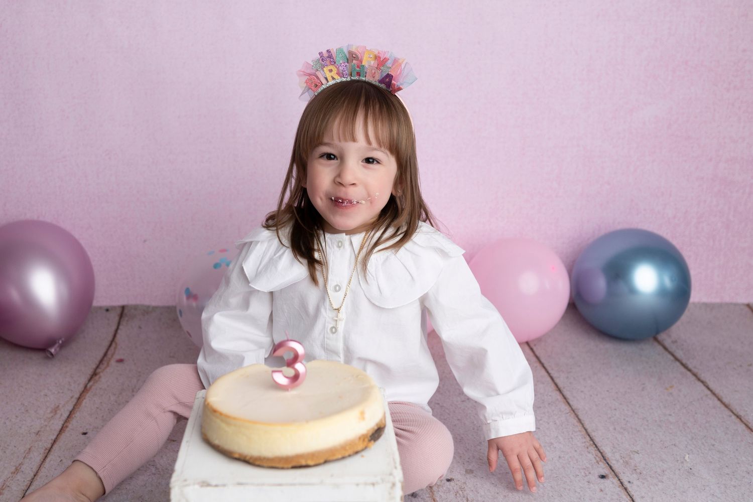 A young girl in a white blouse and birthday crown sits beside a cheesecake with a number 3 candle, surrounded by pastel balloons.