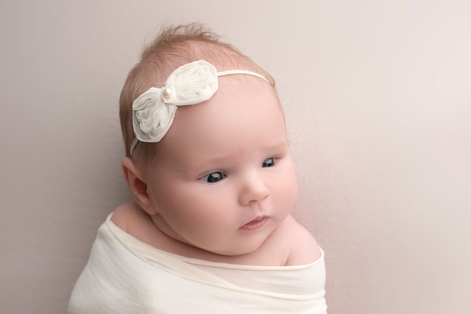 Newborn baby girl wrapped in white swaddle wearing a delicate ivory bow headband against a neutral background.