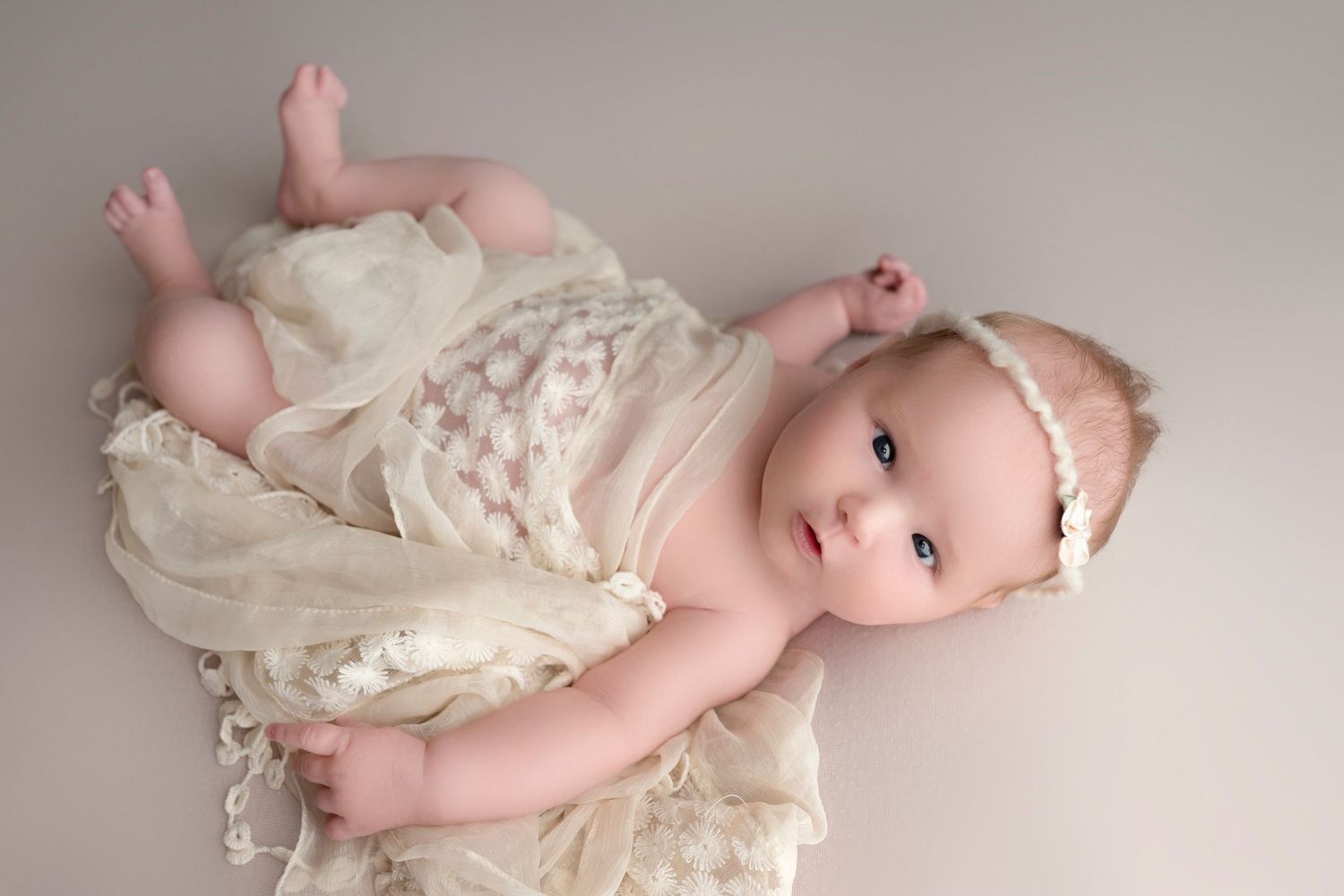 Adorable baby girl in cream lace wrap and floral headband lying on neutral background in newborn portrait.