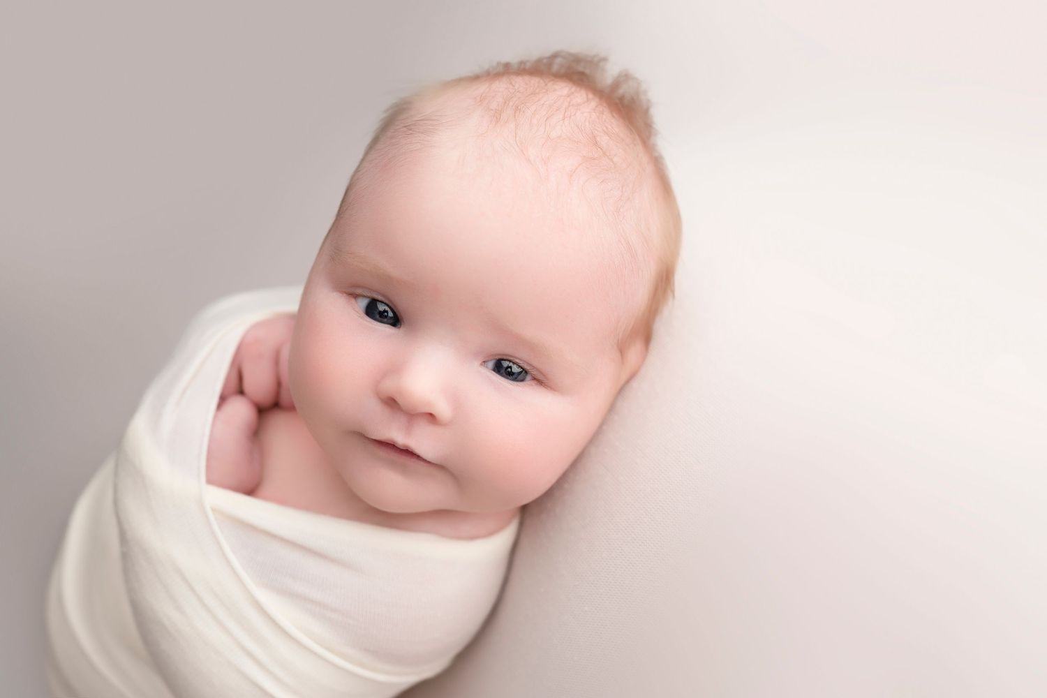Newborn baby swaddled in white wrap with blue eyes on a light gray background.
