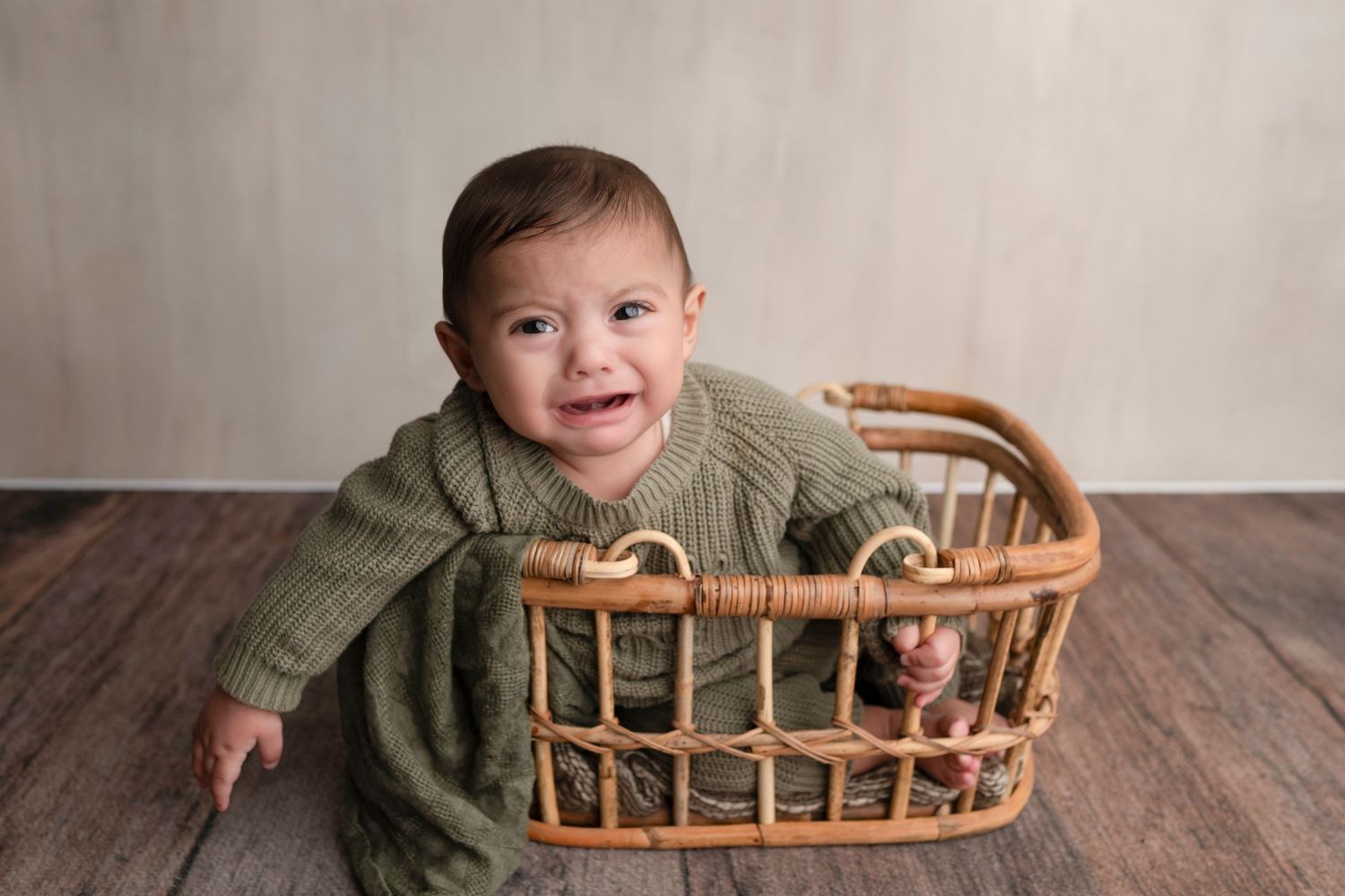 Smiling baby in green knit romper sitting in a wicker basket on a wood floor.