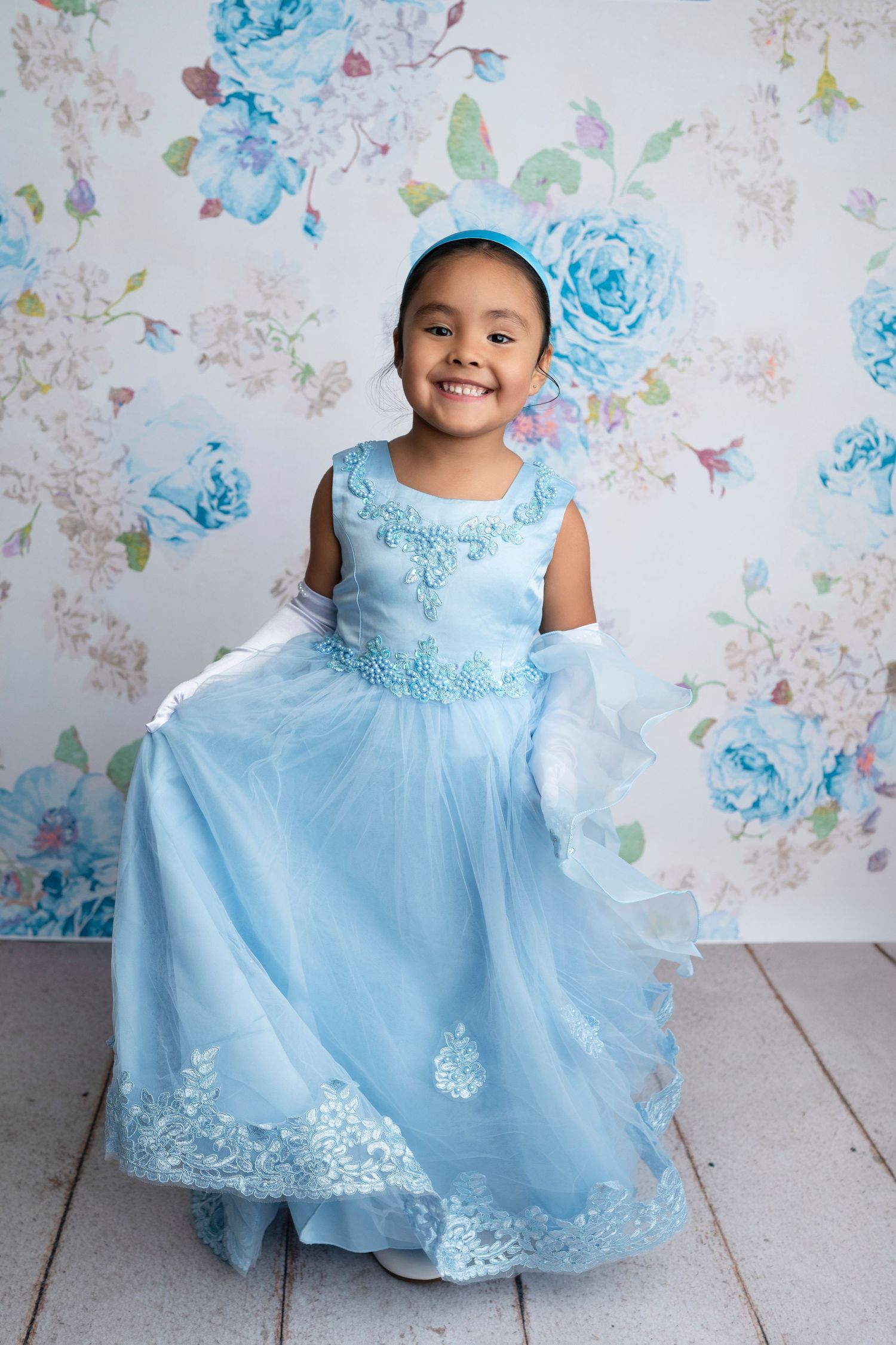 Young girl smiling in a light blue princess gown with lace trim and matching headband against floral backdrop.