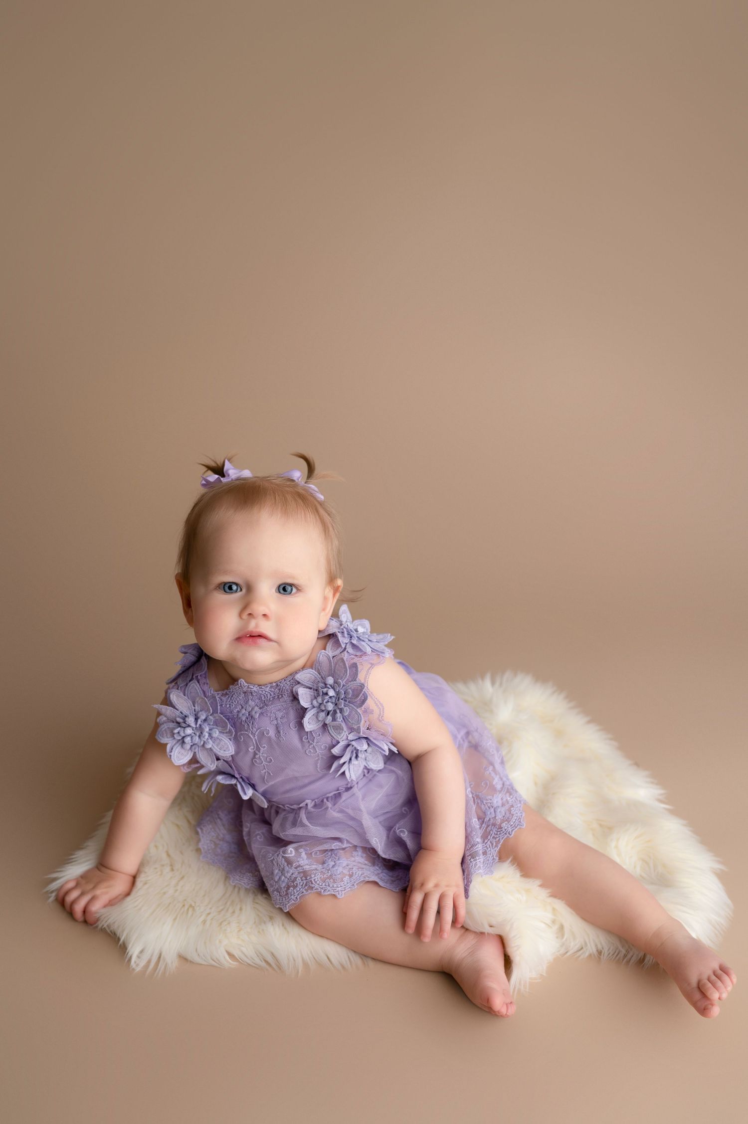 Baby girl in purple lace romper sitting on white fur rug against beige background.