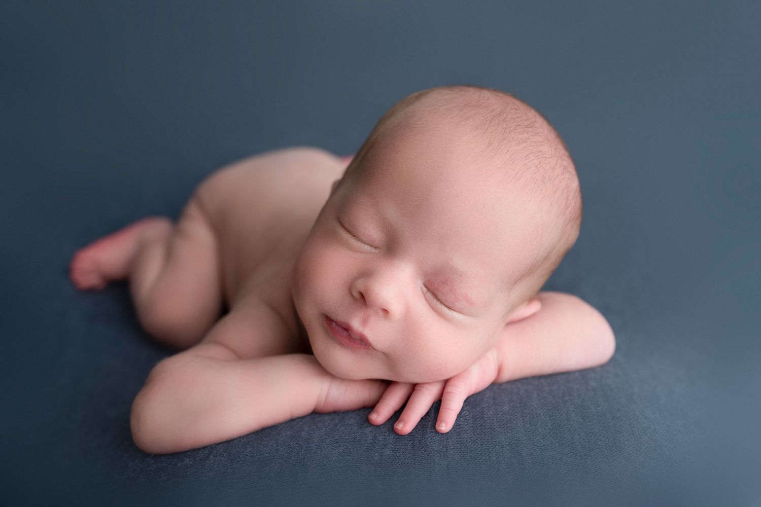 Peaceful newborn baby sleeping on a teal blue blanket with hands tucked under chin in a classic froggy pose.