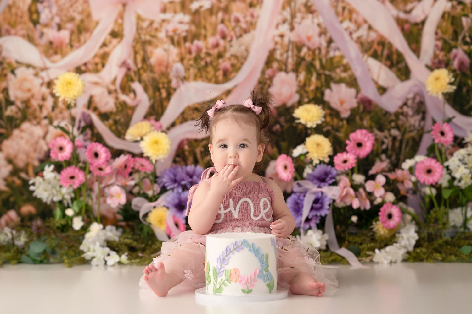 Baby girl in purple outfit smashes first birthday cake surrounded by colorful flowers and pink floral backdrop.
