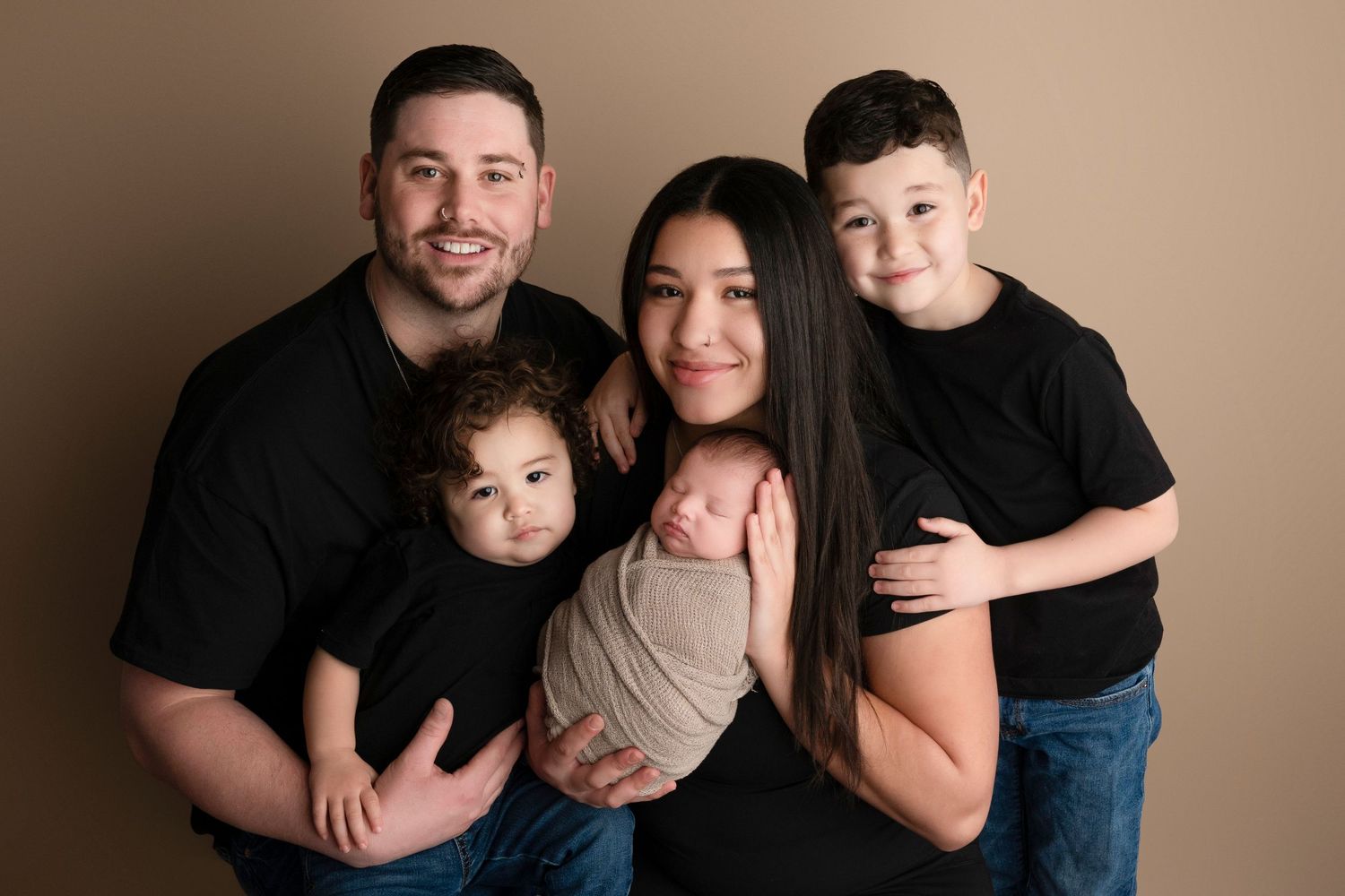 Family of five posing for a newborn portrait session, parents and two young boys in matching black shirts holding swaddled baby.