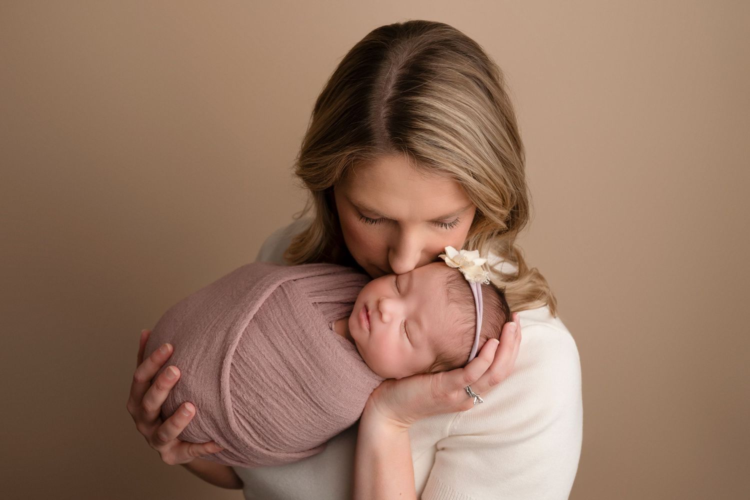 A mother tenderly kisses her swaddled newborn baby wrapped in pink, against a warm beige studio background.