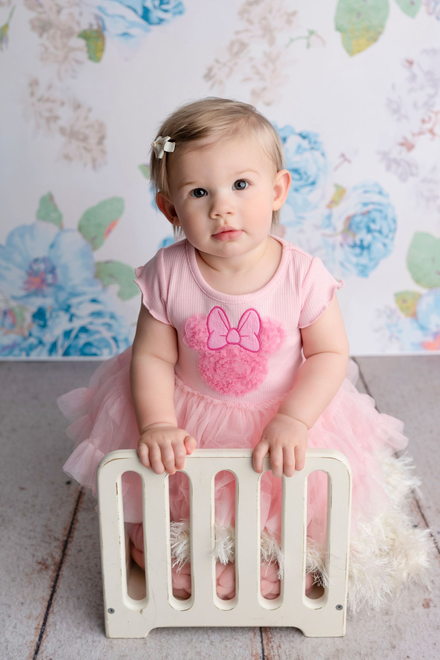 Baby girl in pink Minnie Mouse outfit and tutu skirt sits by white crib prop against floral backdrop.