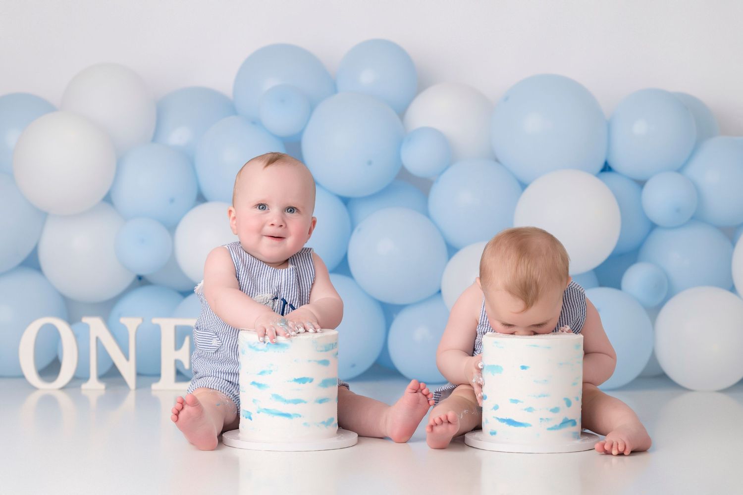 Two babies in blue rompers celebrate their first birthday with white cakes and blue balloon backdrop with ONE sign.