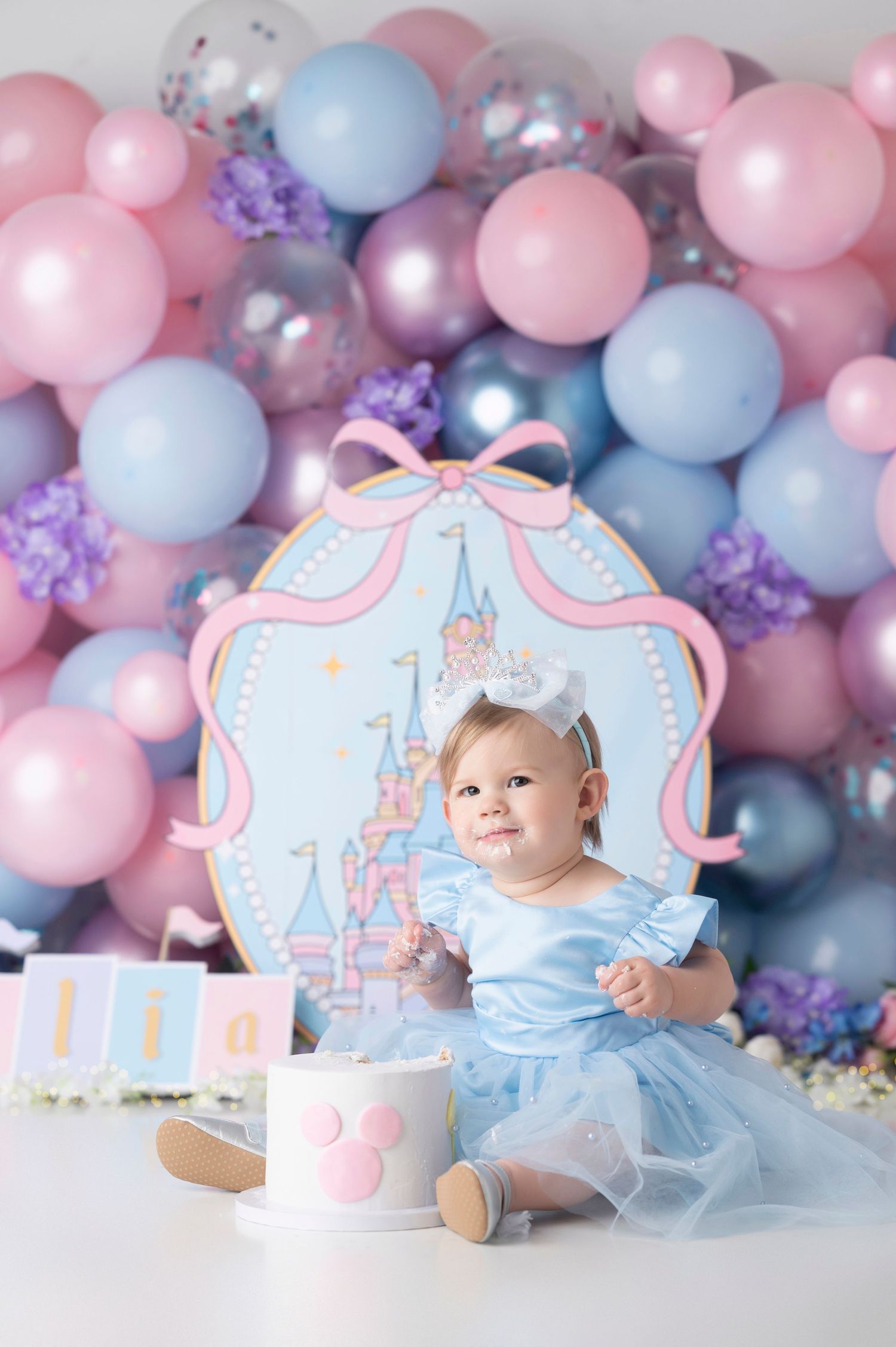 Baby girl in blue dress celebrates 1st birthday with Disney castle backdrop and pink and blue balloon garland.