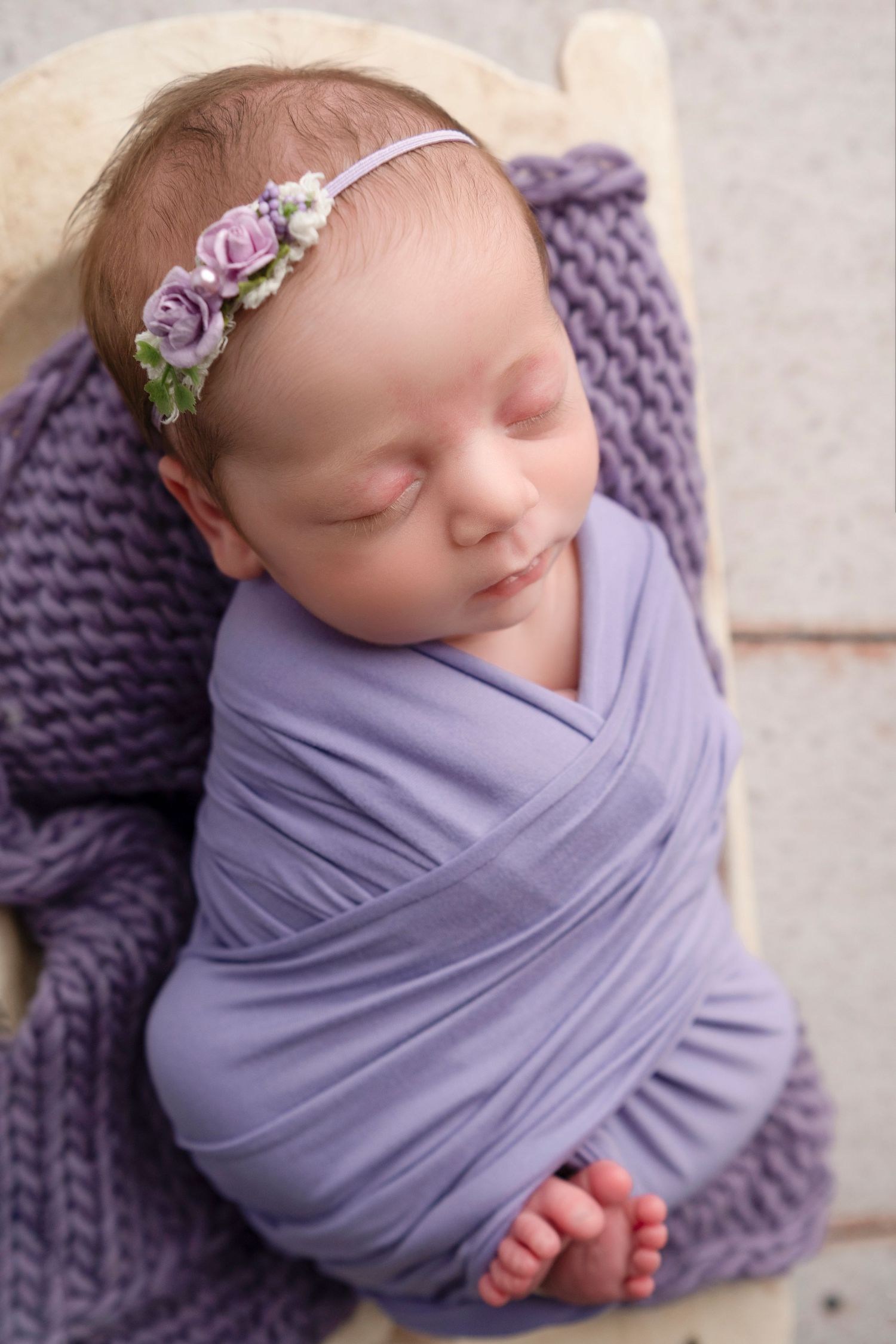 A sleeping newborn wrapped in soft purple fabric with a delicate floral headband rests on a knitted blanket.