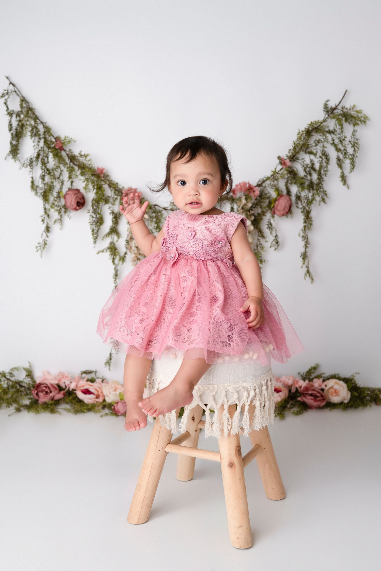 A baby in a pink lace dress sits on a rustic wooden stool against a white backdrop with evergreen branches.