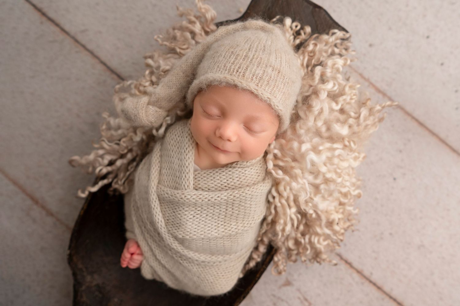 A sleeping newborn baby wrapped in beige knit swaddle lies on soft textured blanket in professional studio setting.