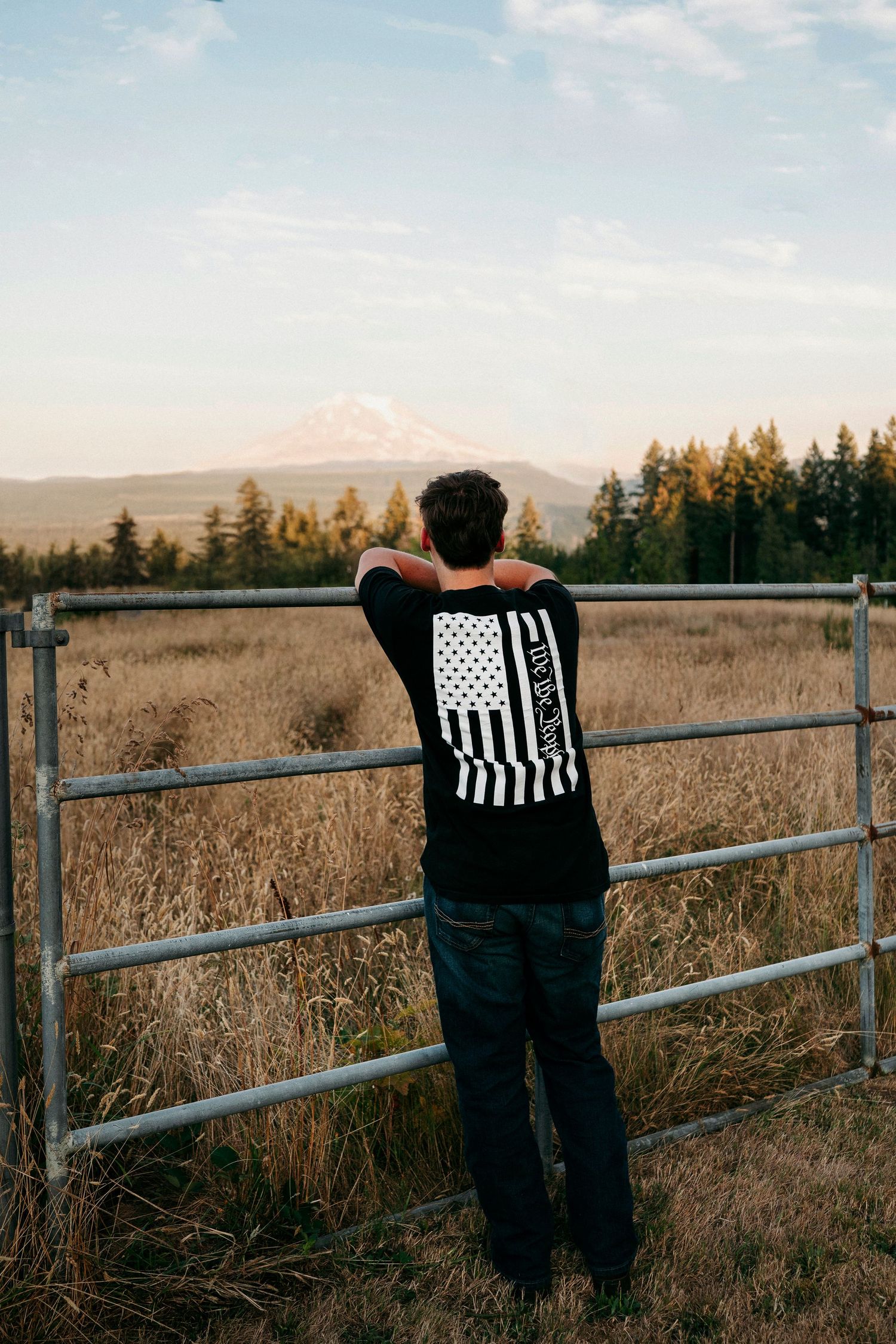 Figure leaning against wooden fence posts overlooking dry grassland with evergreen forest in background.