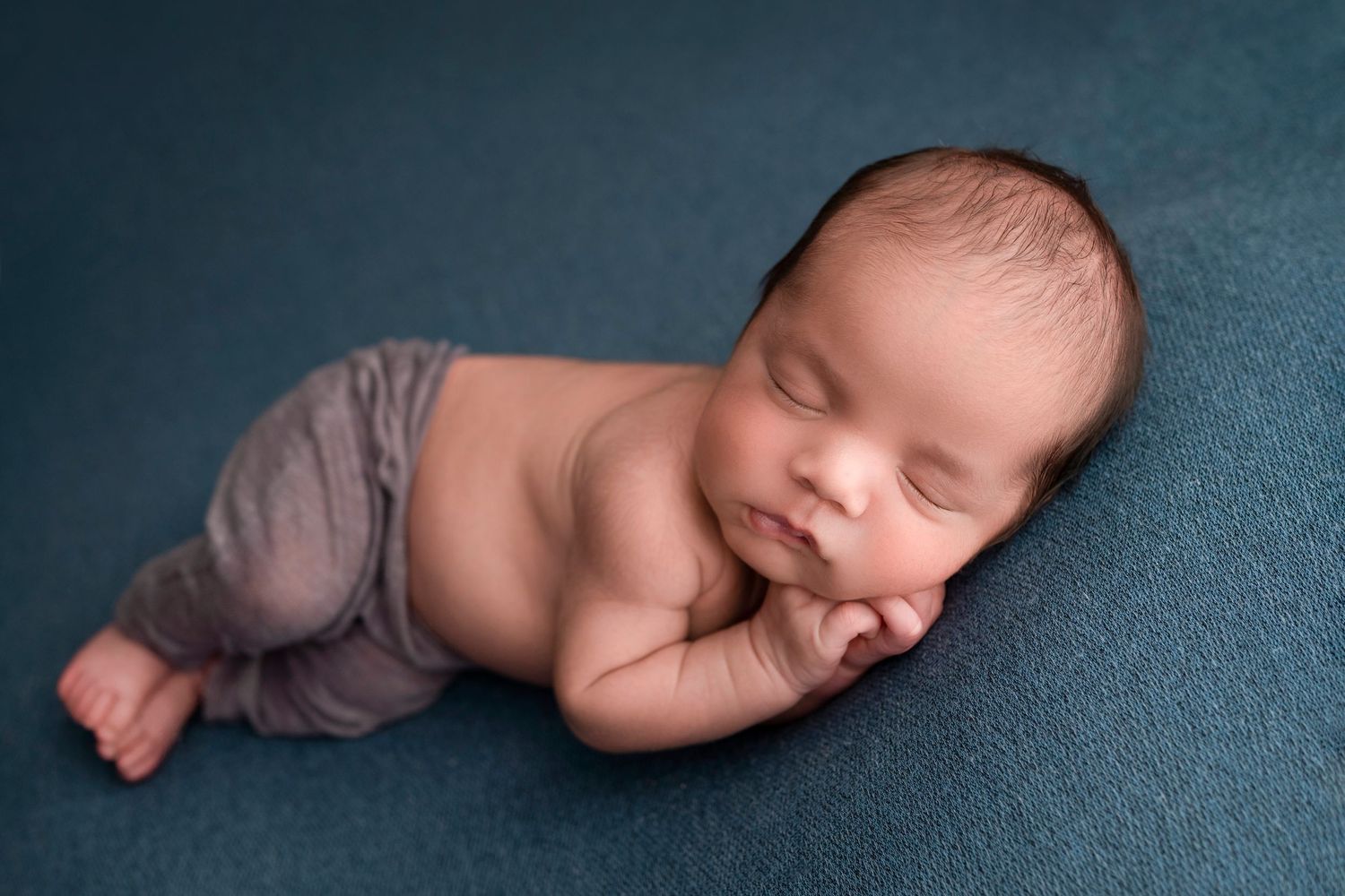 Peaceful newborn baby sleeping on teal fabric, wearing gray pants, curled in a natural pose for a professional photo.