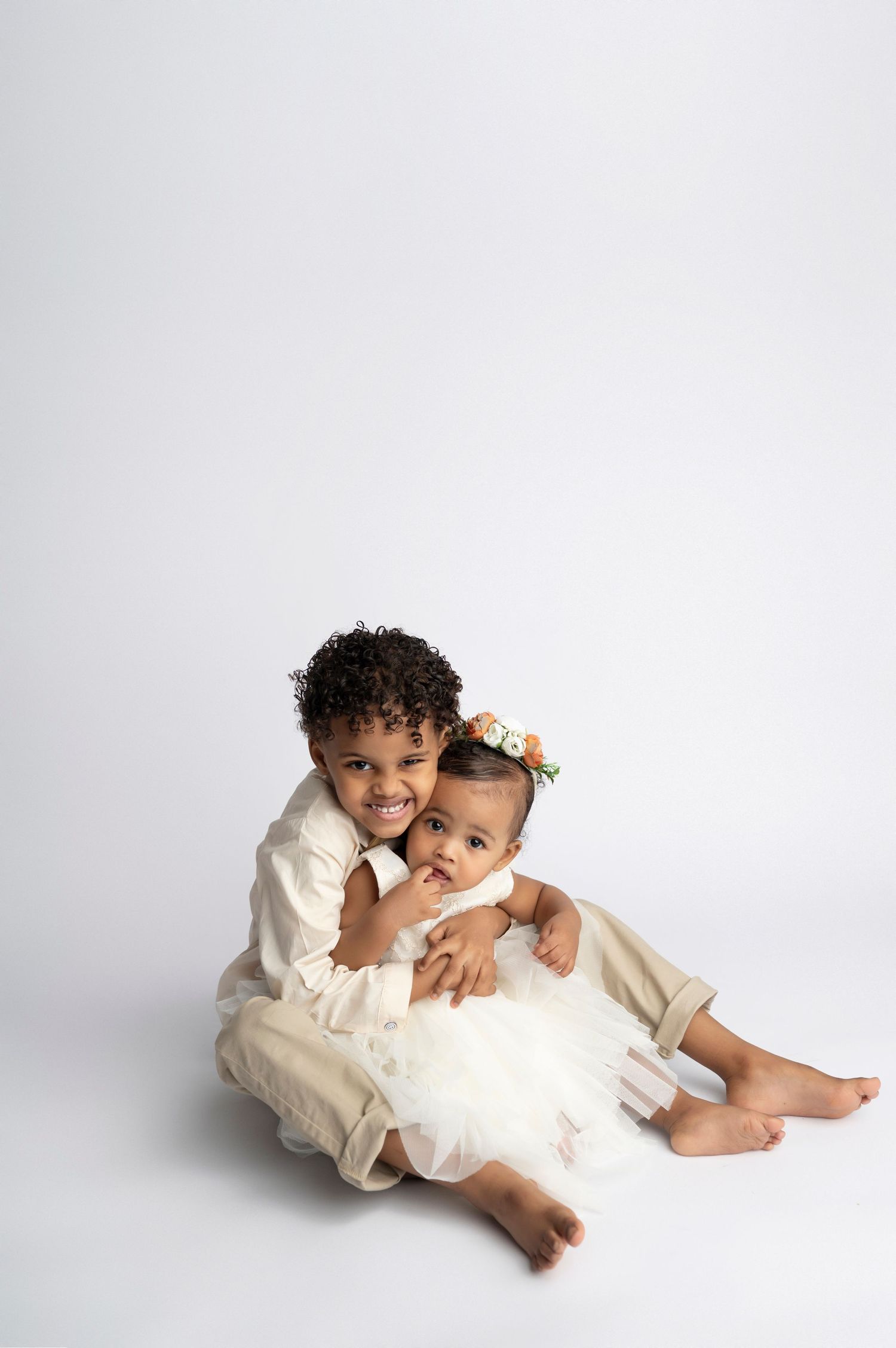 Two young children in cream outfits sit together, older child hugging baby girl wearing floral crown, on white background.