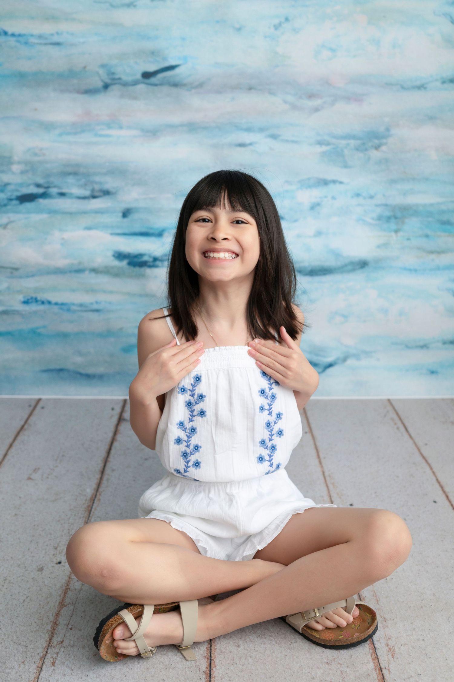 Smiling girl in white embroidered romper sitting cross-legged on floor with blue watercolor backdrop.