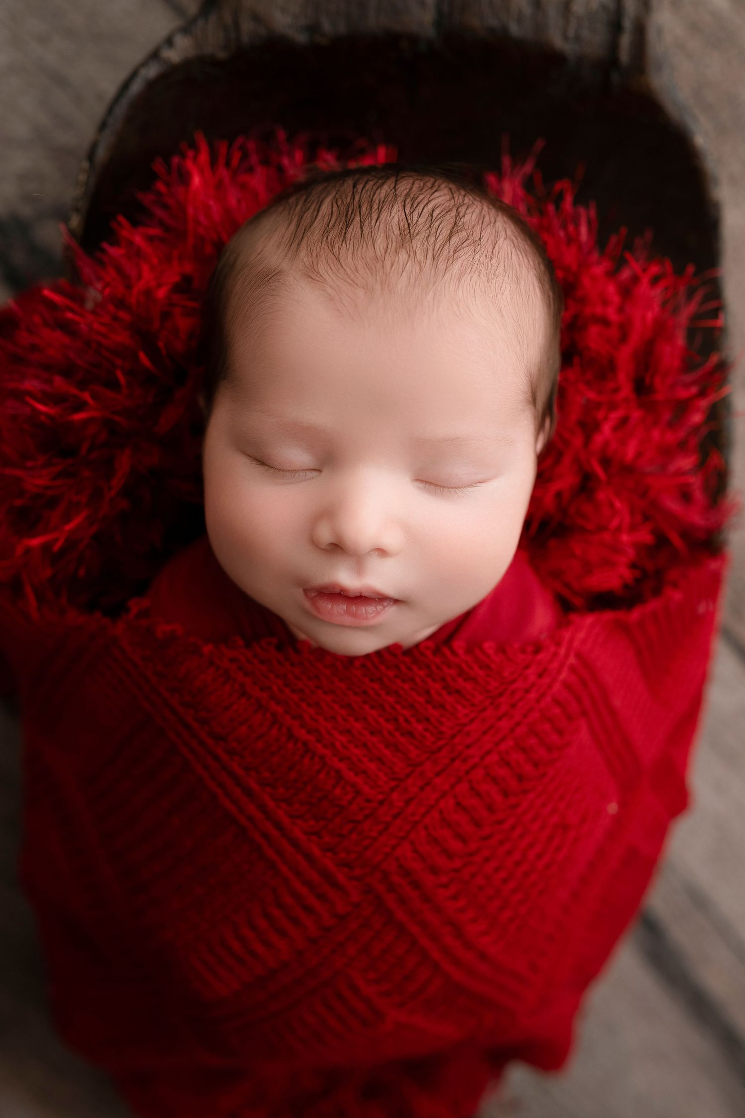 Newborn baby sleeping peacefully swaddled in a red knit wrap with fluffy red trim inside a wooden bowl.