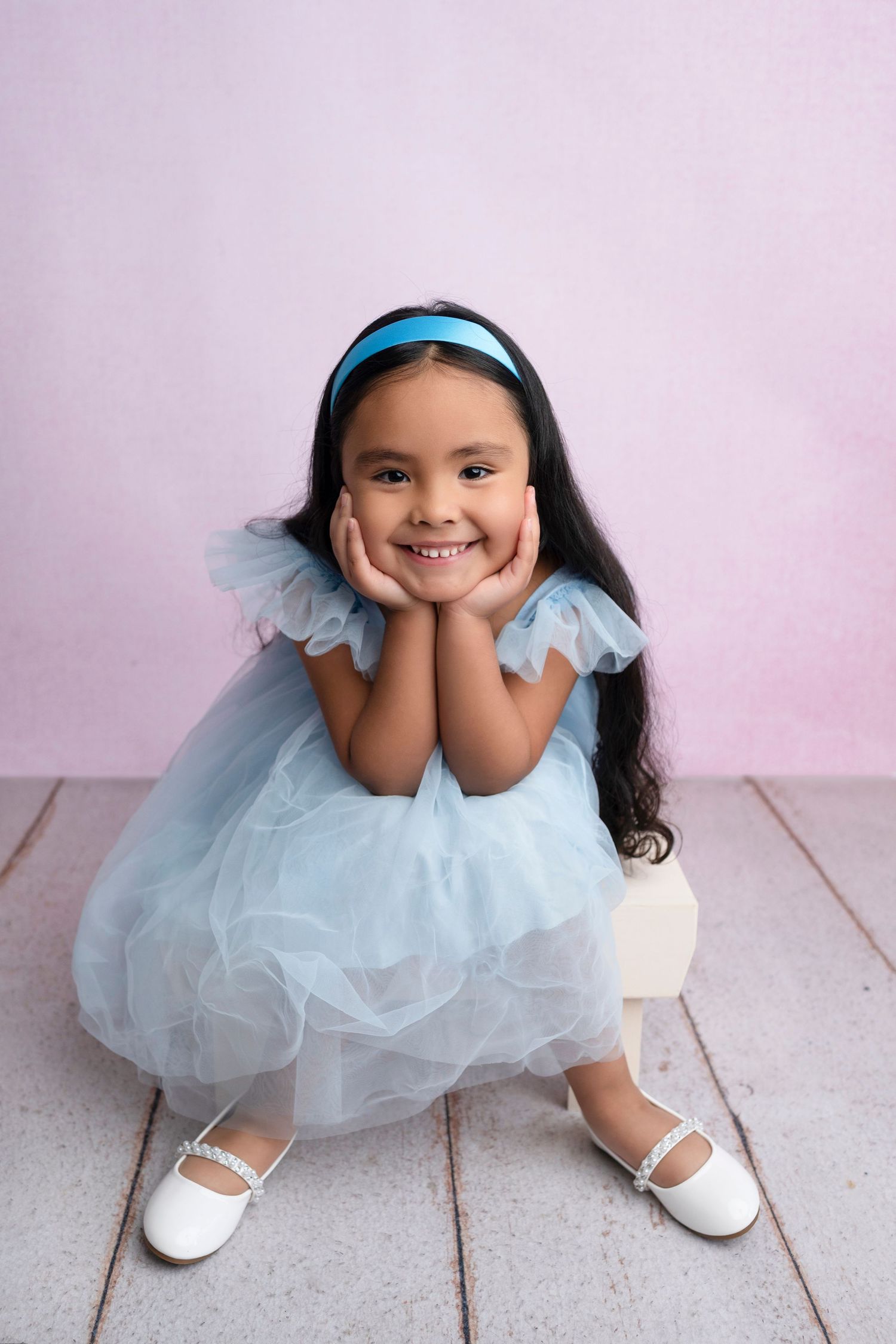 Young girl in blue tulle dress and headband smiles with hands on cheeks, sitting on white wooden floor.