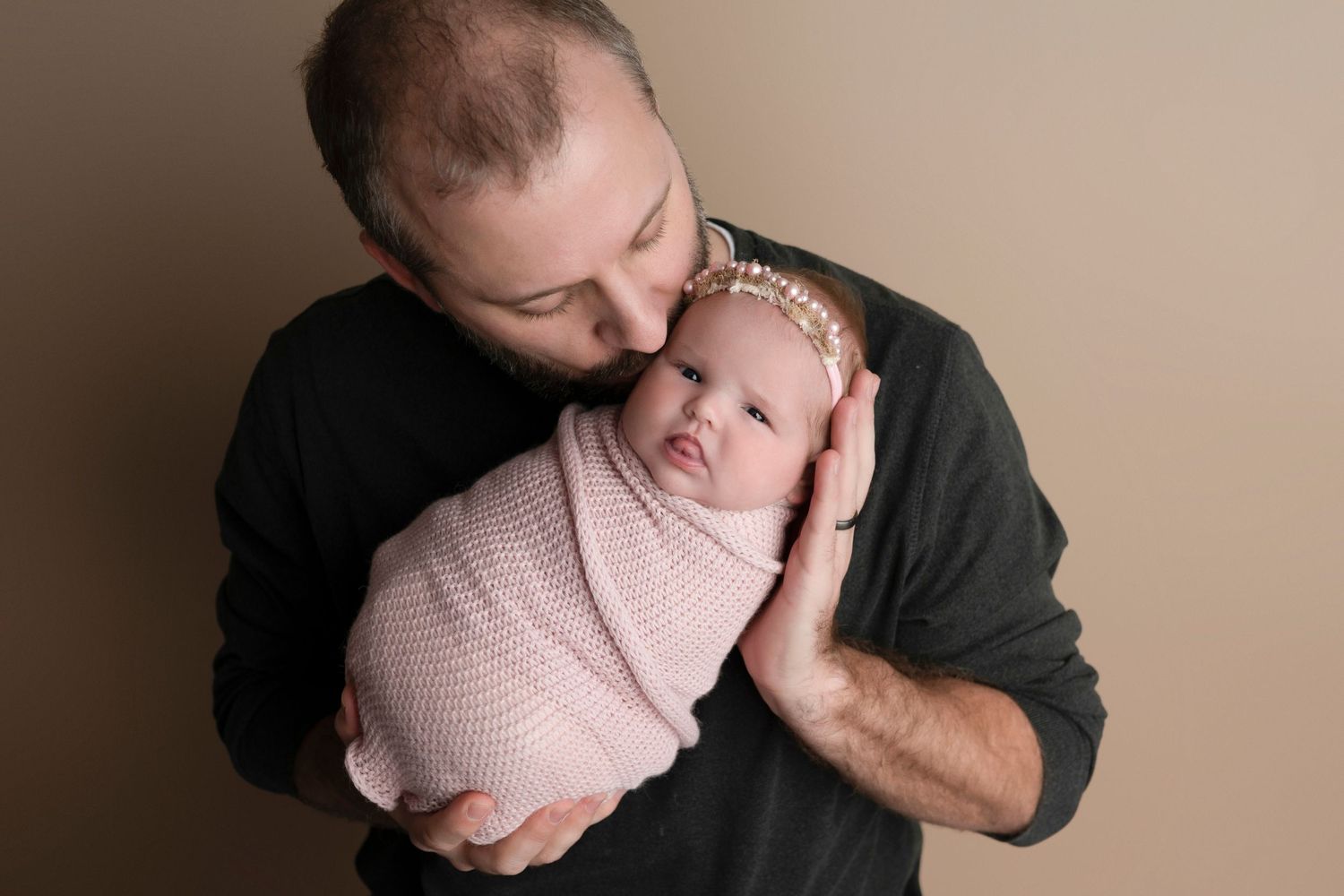 A father in a black shirt tenderly kisses his swaddled newborn baby girl wearing a pink headband.