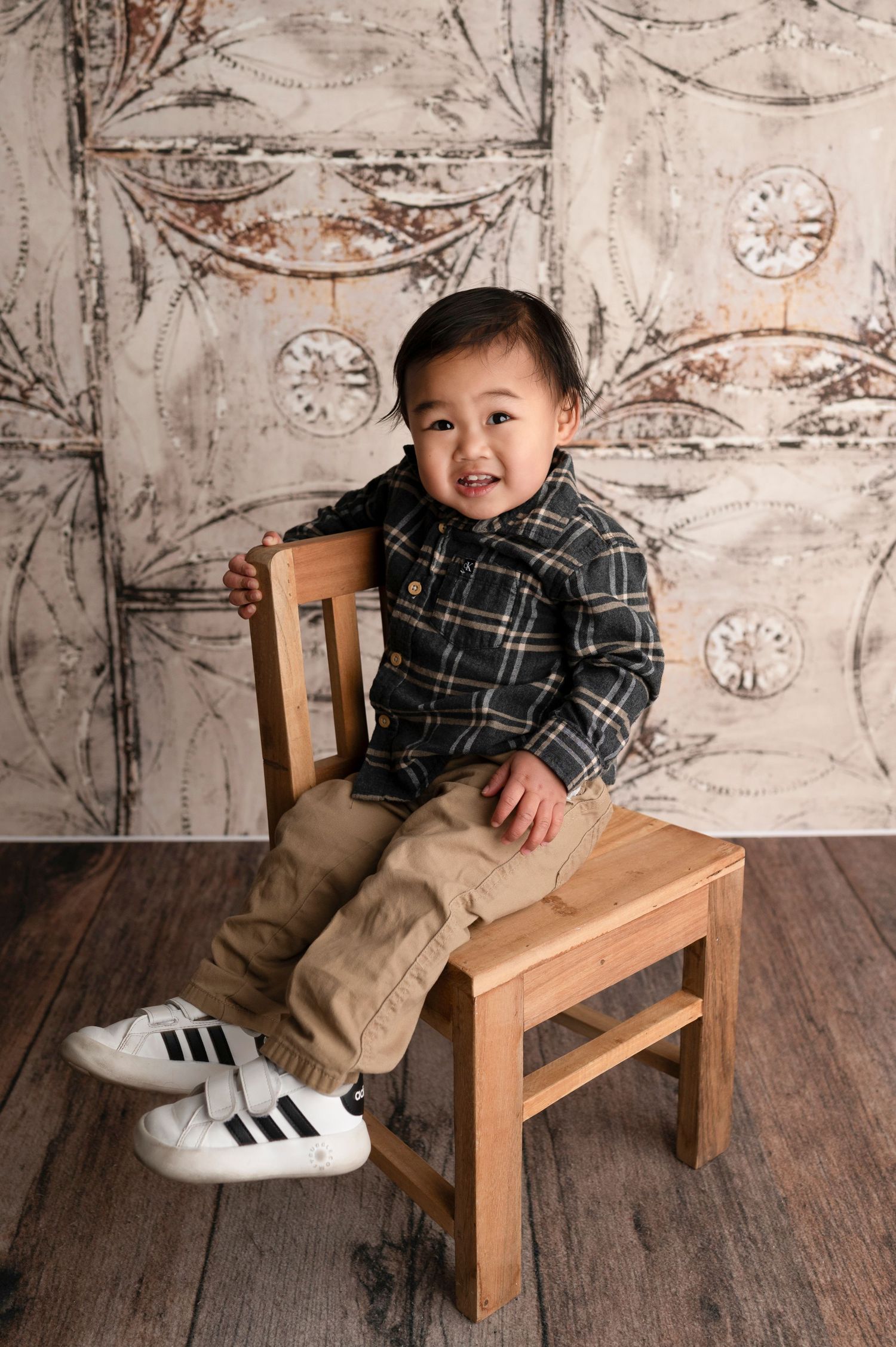 Smiling toddler boy in plaid shirt and khaki pants sitting on a small wooden chair against a vintage backdrop.