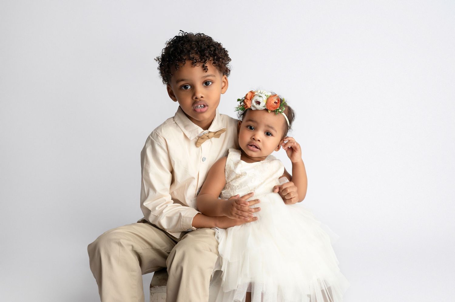 Two young children in formal attire pose together, boy in tan suit and baby girl in white dress with floral crown.