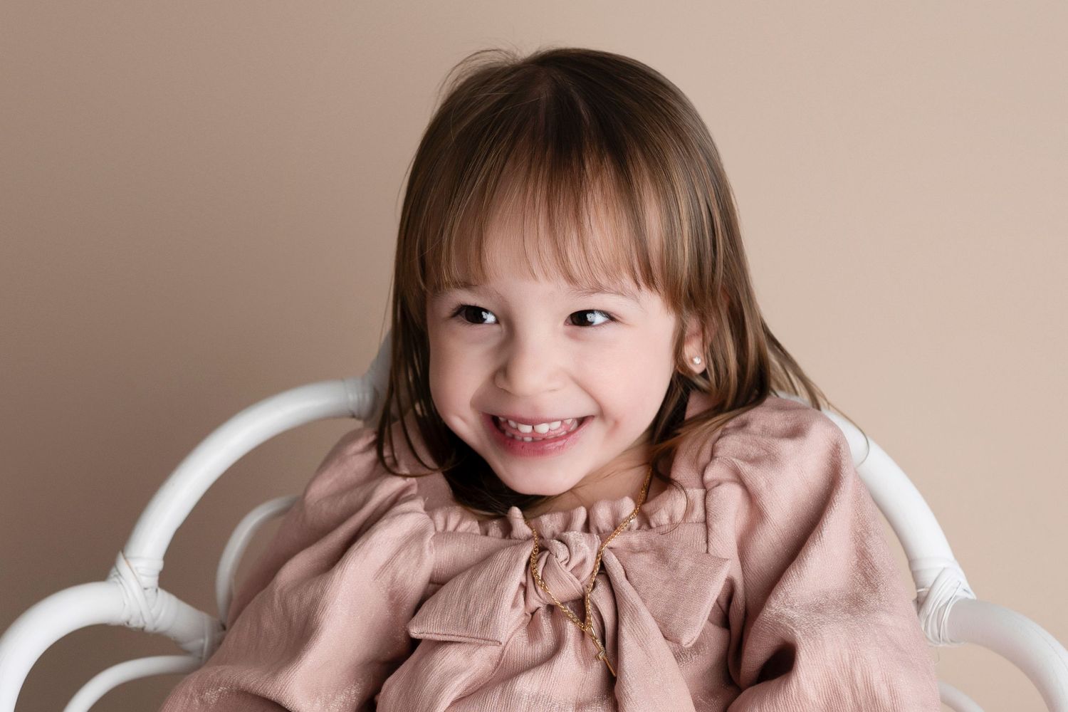 Smiling toddler girl with brown hair wearing a pink linen dress, seated in a white chair against a beige background.