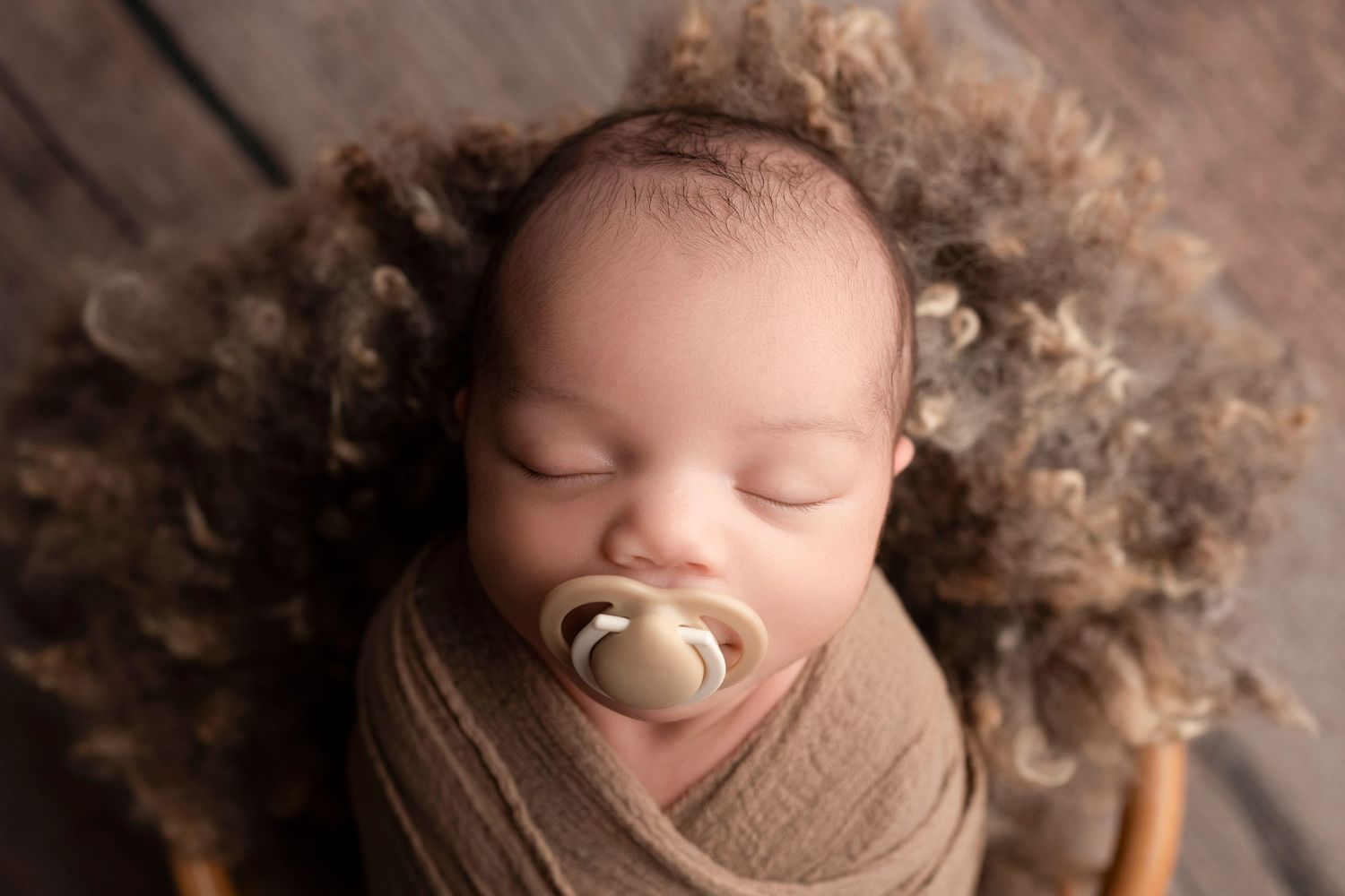 A sleeping newborn baby swaddled in brown fabric with a pacifier during a professional photography session.