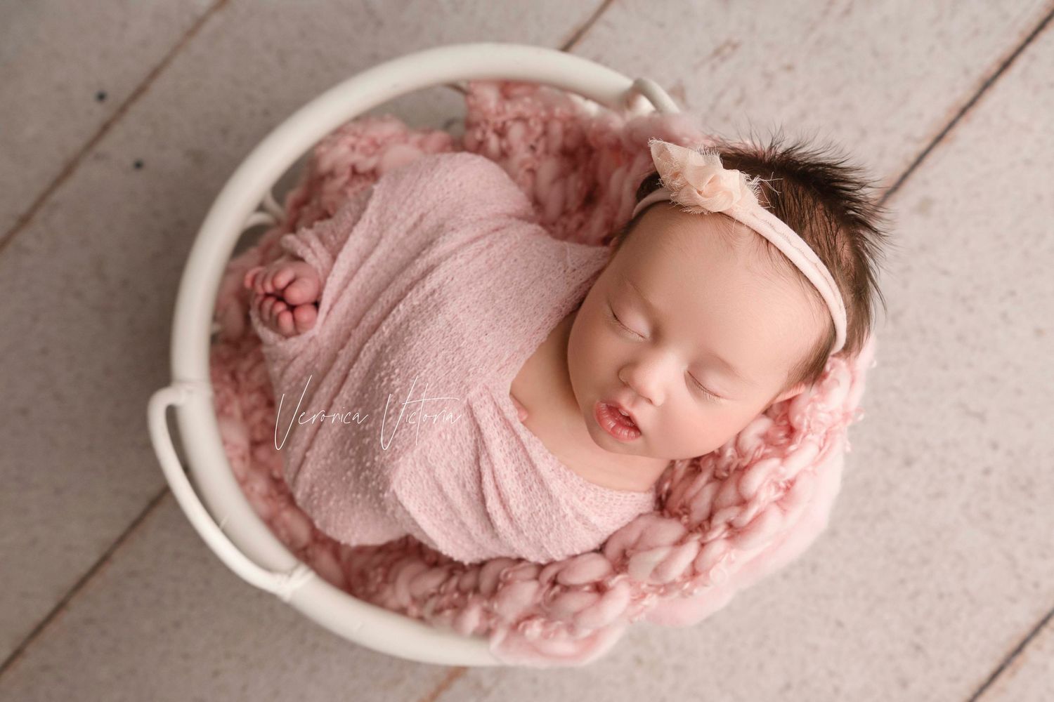 Newborn baby sleeping peacefully in pink swaddle and white bowl on wooden floor.
